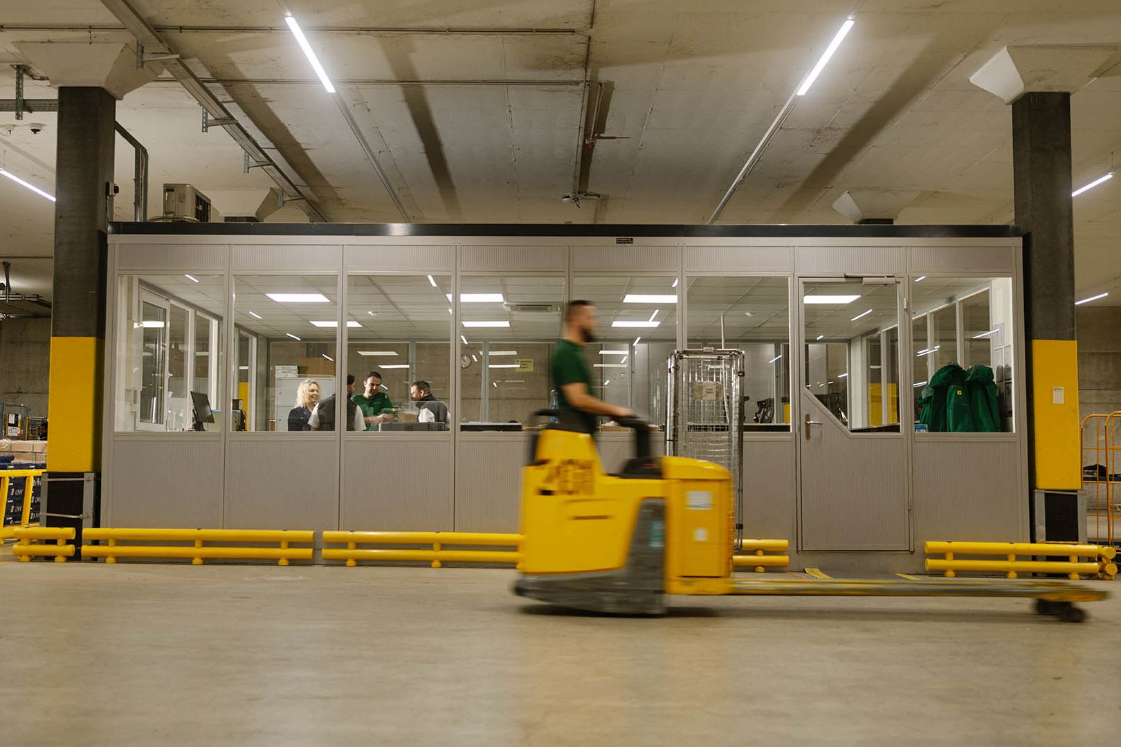 Dans un entrepôt, un employé de Swiss Post Cargo passe avec un transpalette devant une pièce vitrée.