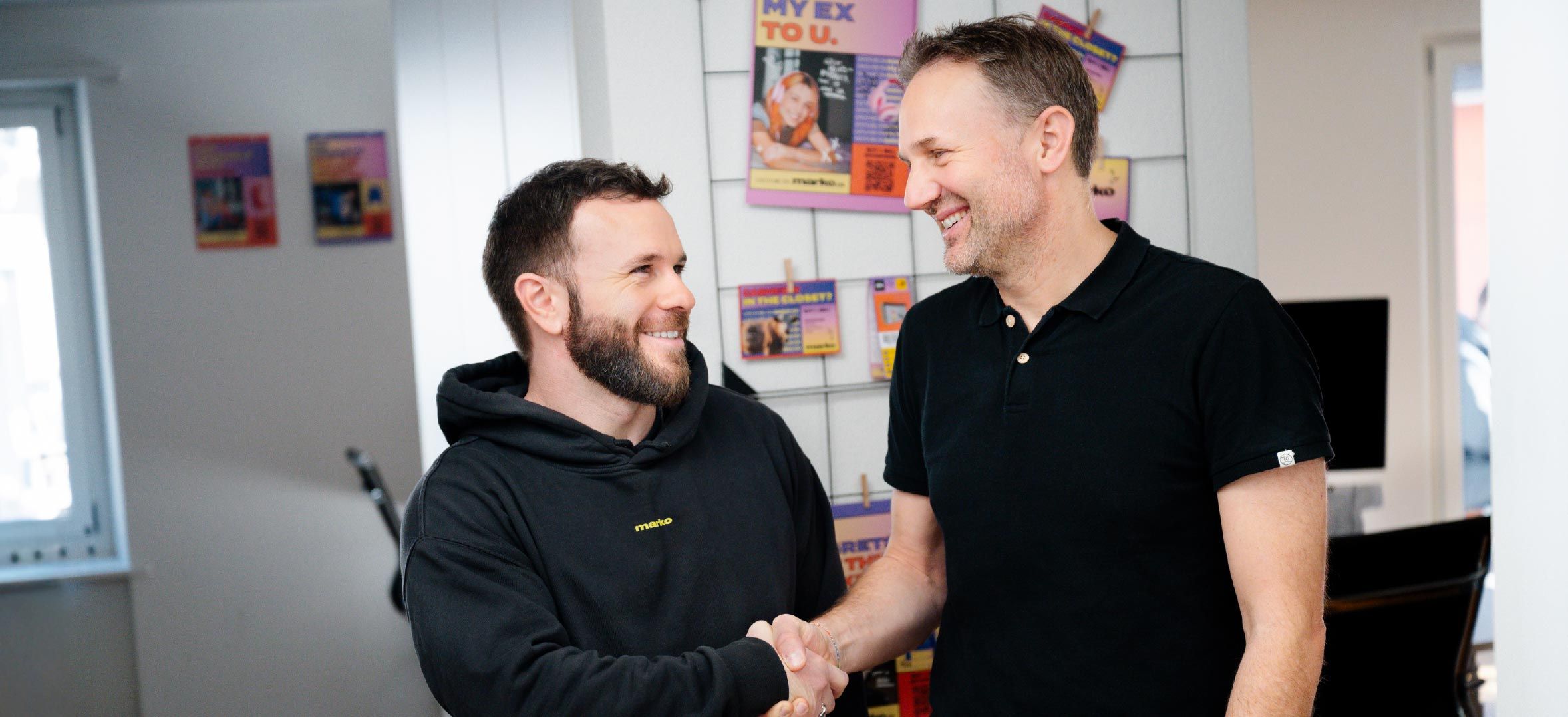 Due persone si stringono la mano in un ambiente interno. Sullo sfondo scaffali con opuscoli colorati e una finestra.