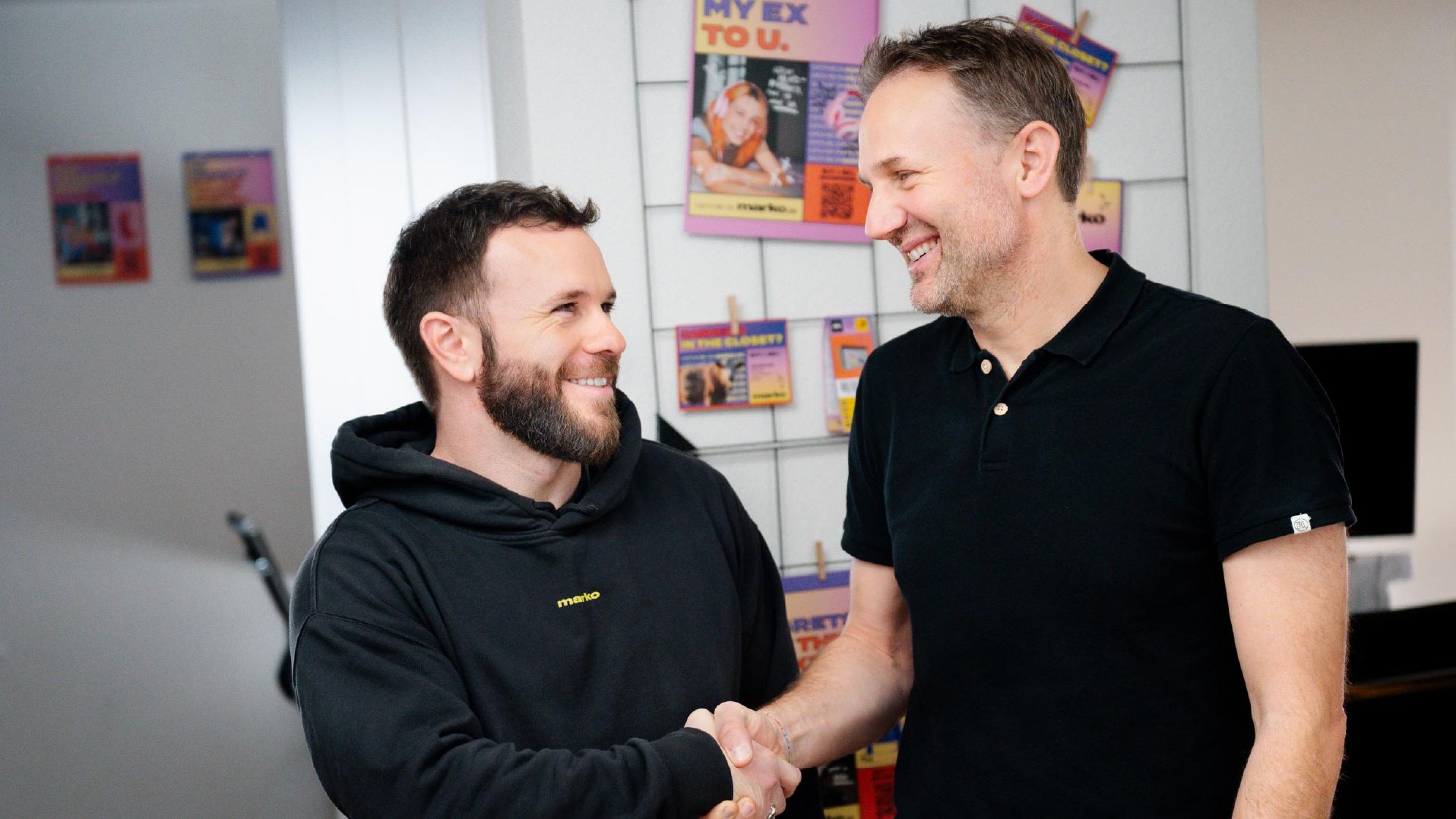 Due persone si stringono la mano in un ambiente interno. Sullo sfondo scaffali con opuscoli colorati e una finestra.