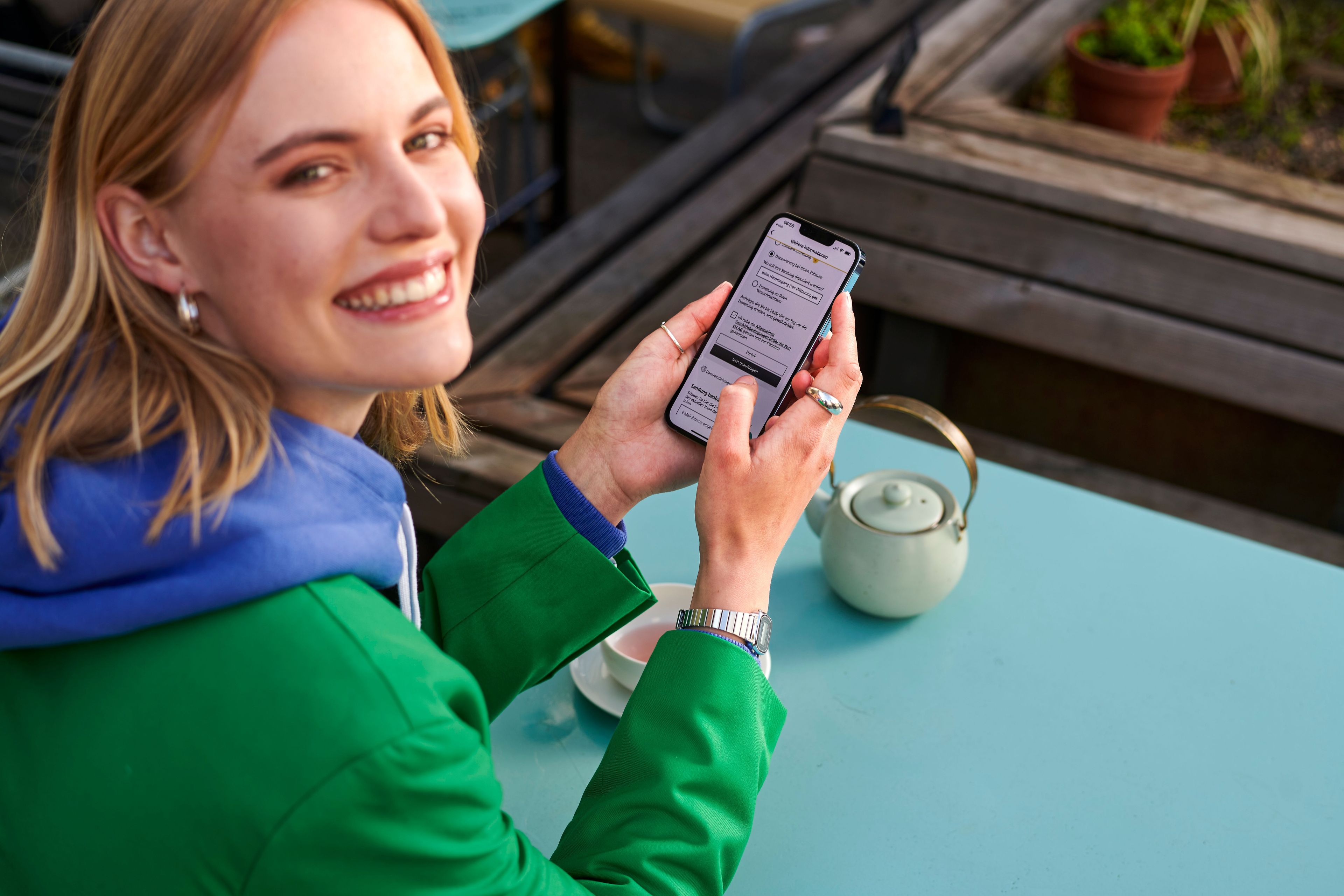 Person sitzt an einem Tisch im Freien und hält ein Smartphone in der Hand. Auf dem Tisch steht eine helle Teekanne. Die Person betrachtet eine geöffnete App oder Webseite auf dem Bildschirm.