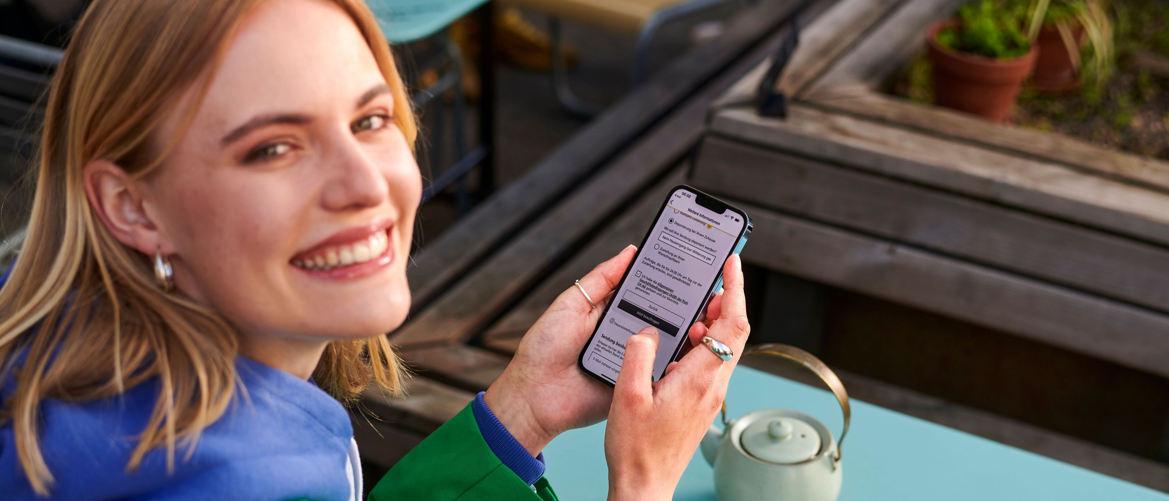 Person sitzt an einem Tisch im Freien und hält ein Smartphone in der Hand. Auf dem Tisch steht eine helle Teekanne. Die Person betrachtet eine geöffnete App oder Webseite auf dem Bildschirm.