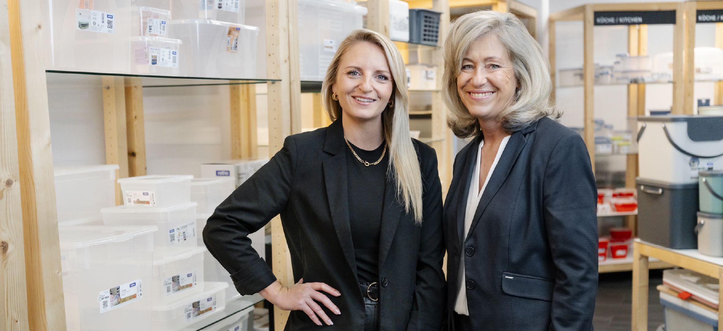 Zwei Personen stehen in einem Raum mit Regalen voller transparenter Kunststoffboxen und Aufbewahrungsbehälter. Im Hintergrund sind weitere Regale mit verschiedenen Organisationsprodukten sichtbar.