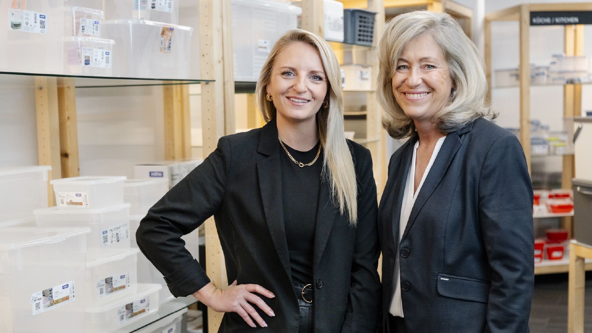 Zwei Personen stehen in einem Raum mit Regalen voller transparenter Kunststoffboxen und Aufbewahrungsbehälter. Im Hintergrund sind weitere Regale mit verschiedenen Organisationsprodukten sichtbar.