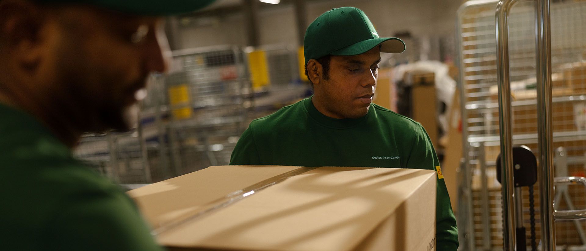 Zwei Swiss Post Cargo Mitarbeitende laden Pakete von einem Fliessband auf einen Rollwagen.