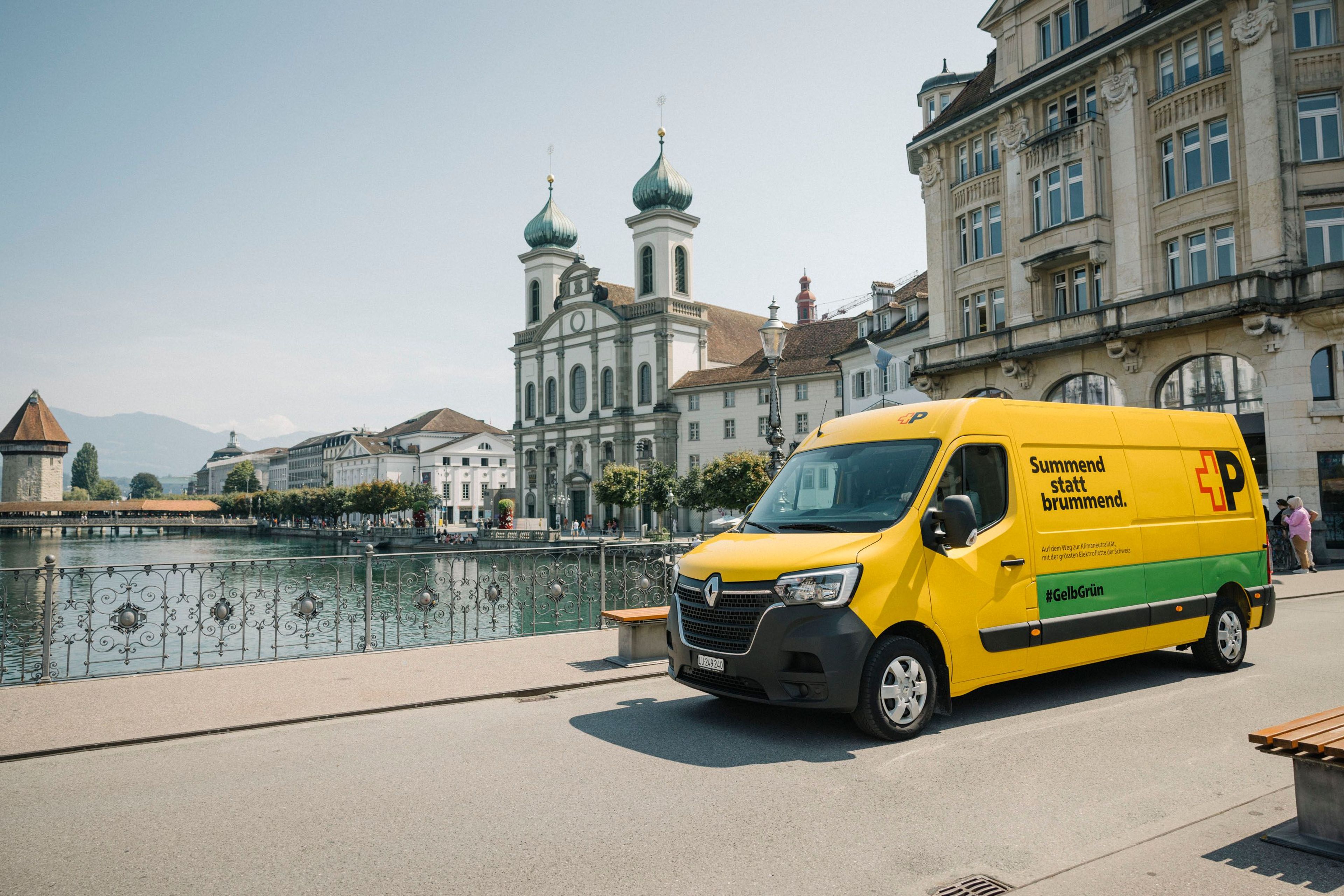 Un furgone giallo della Posta Svizzera è parcheggiato su una passeggiata lungo il fiume a Lucerna. Sullo sfondo la chiesa dei Gesuiti, edifici storici e la Reuss. Testo sul veicolo: «Summend statt brummend.».