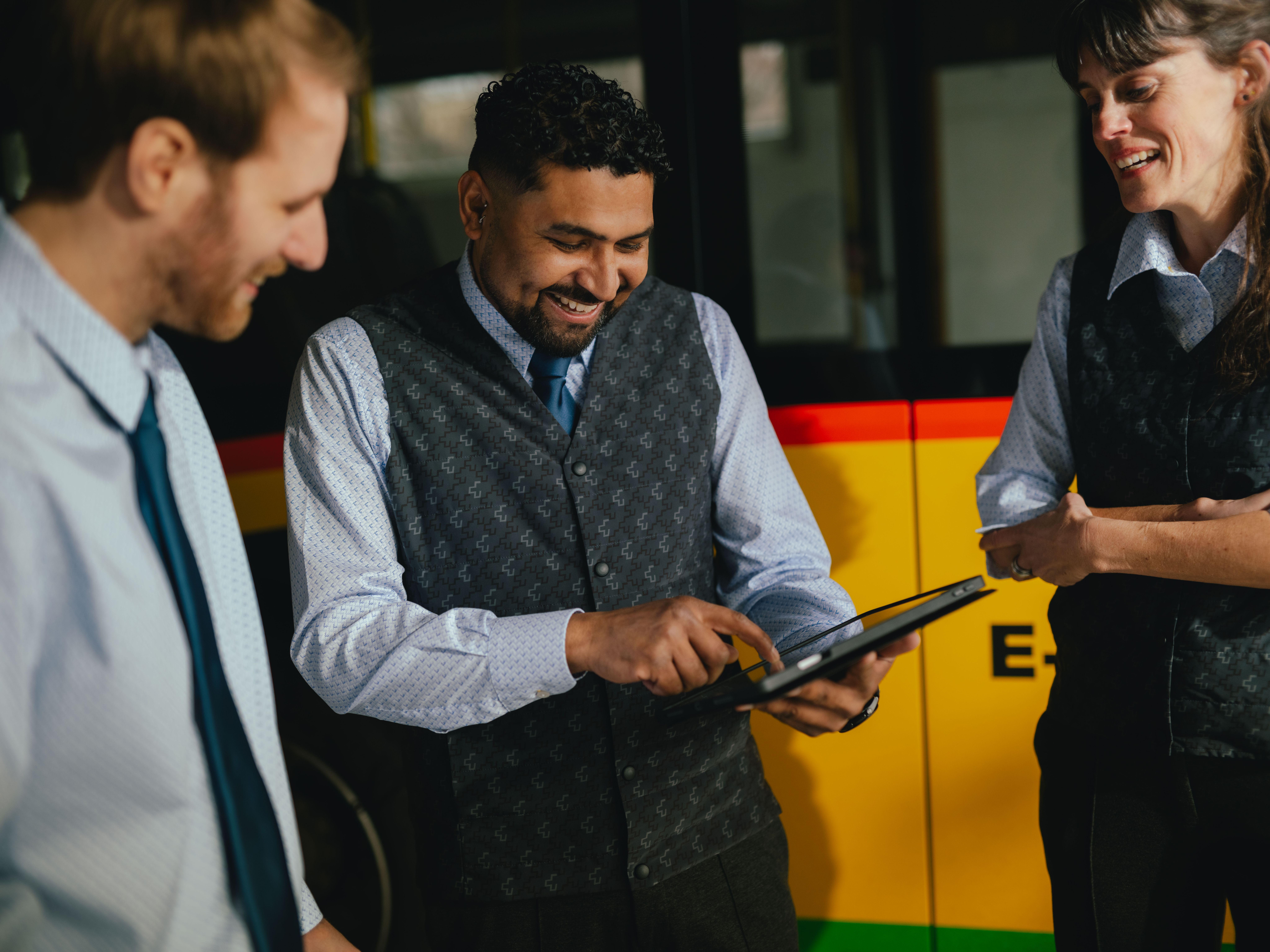 Three people are standing in a hall in front of a yellow and black vehicle. One person is holding a tablet and pointing at it, while the other two stand nearby observing. All are wearing formal clothing, and the side of the vehicle with coloured stripes and text is visible in the background.