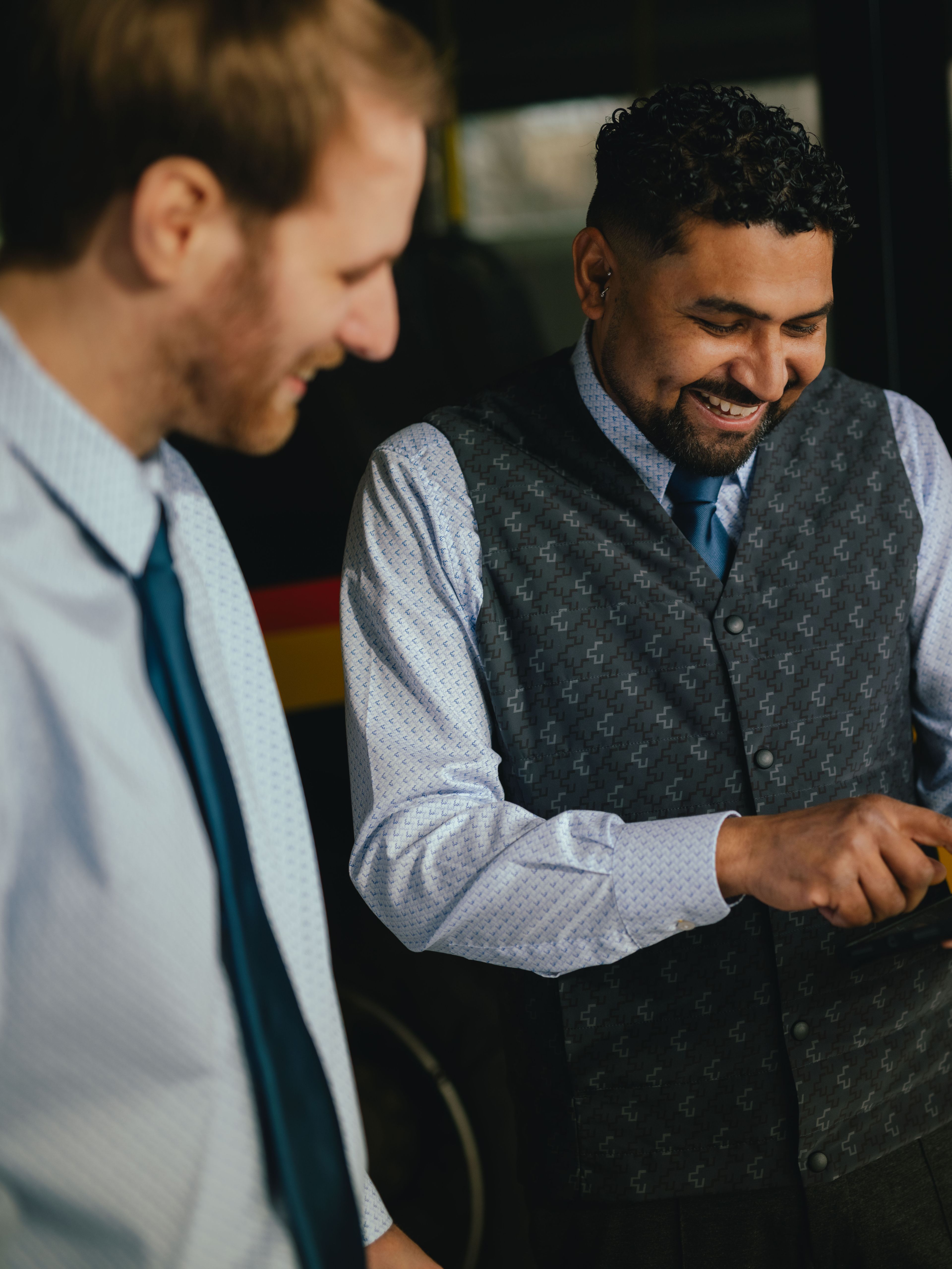 Three people are standing in a hall in front of a yellow and black vehicle. One person is holding a tablet and pointing at it, while the other two stand nearby observing. All are wearing formal clothing, and the side of the vehicle with coloured stripes and text is visible in the background.