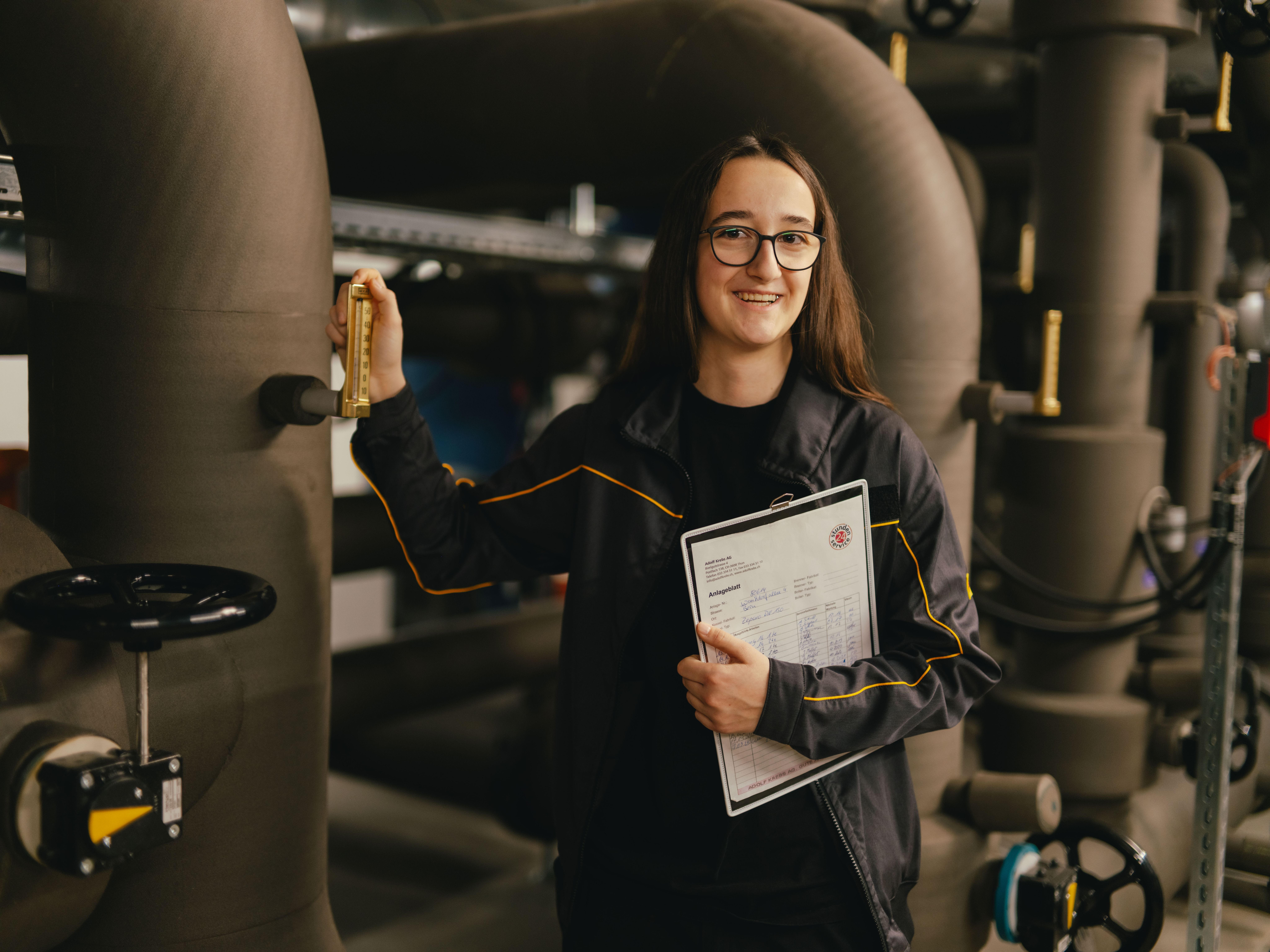 Una persona si trova in un impianto tecnico circondata da grandi tubazioni e tiene una cartellina con documenti tecnici. Con l’altra mano aziona una leva metallica su una delle tubature. L’ambiente è industriale, con valvole, tubi e illuminazione soffusa.