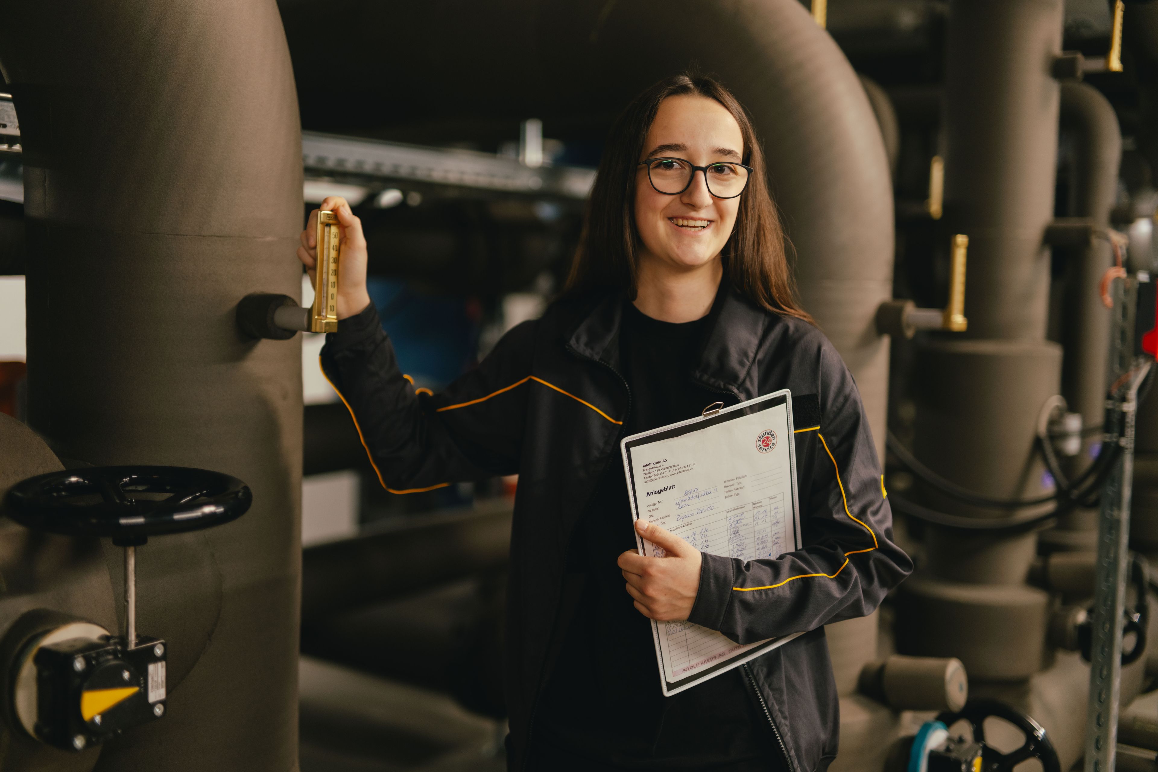 Una persona si trova in un impianto tecnico circondata da grandi tubazioni e tiene una cartellina con documenti tecnici. Con l’altra mano aziona una leva metallica su una delle tubature. L’ambiente è industriale, con valvole, tubi e illuminazione soffusa.