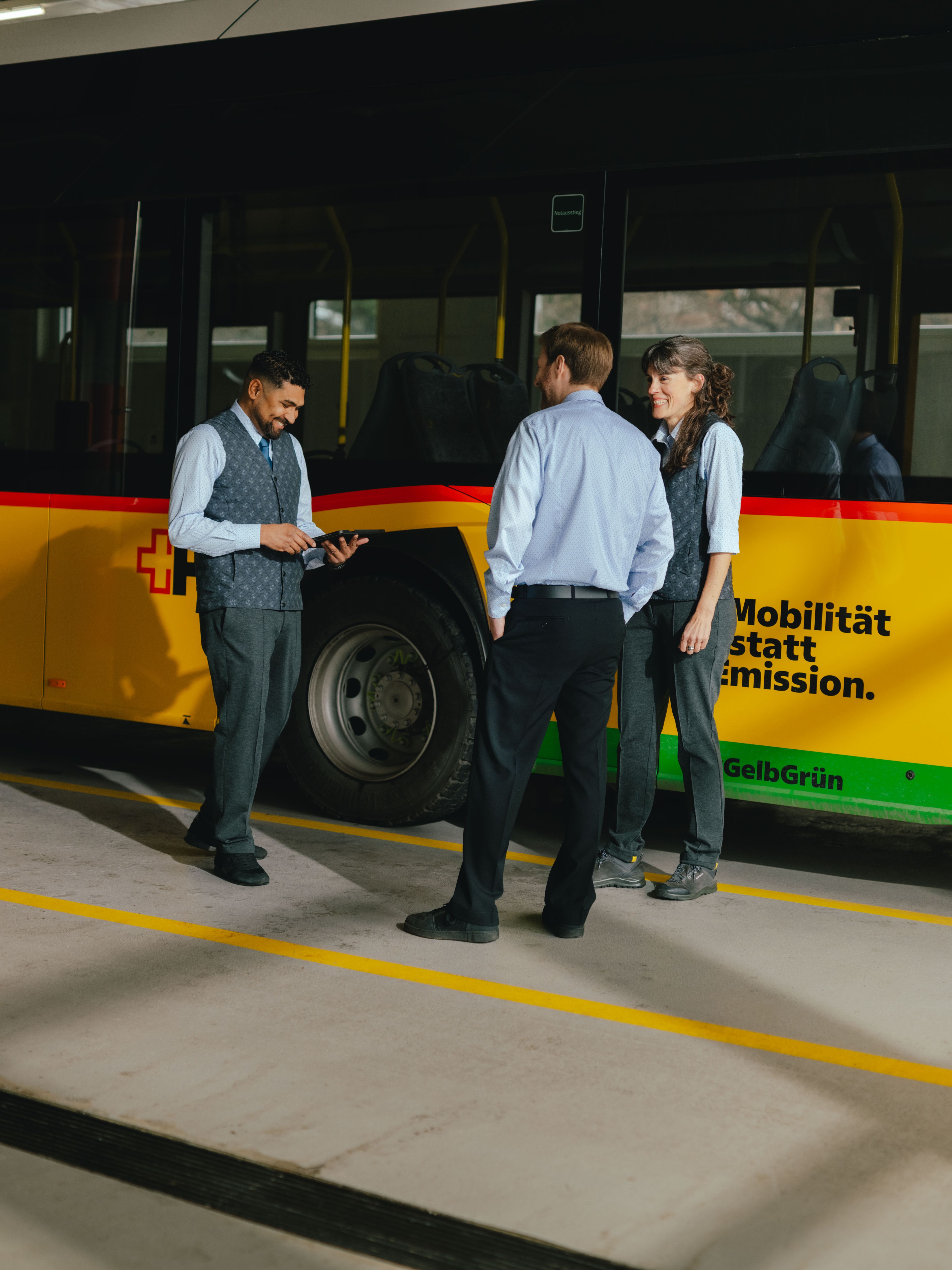 Three people are standing in front of a yellow and black vehicle. One individual is holding a tablet and pointing at the screen, while the other two stand nearby observing. All are wearing formal clothing, such as shirts and vests. The background shows the side of the vehicle with coloured stripes.