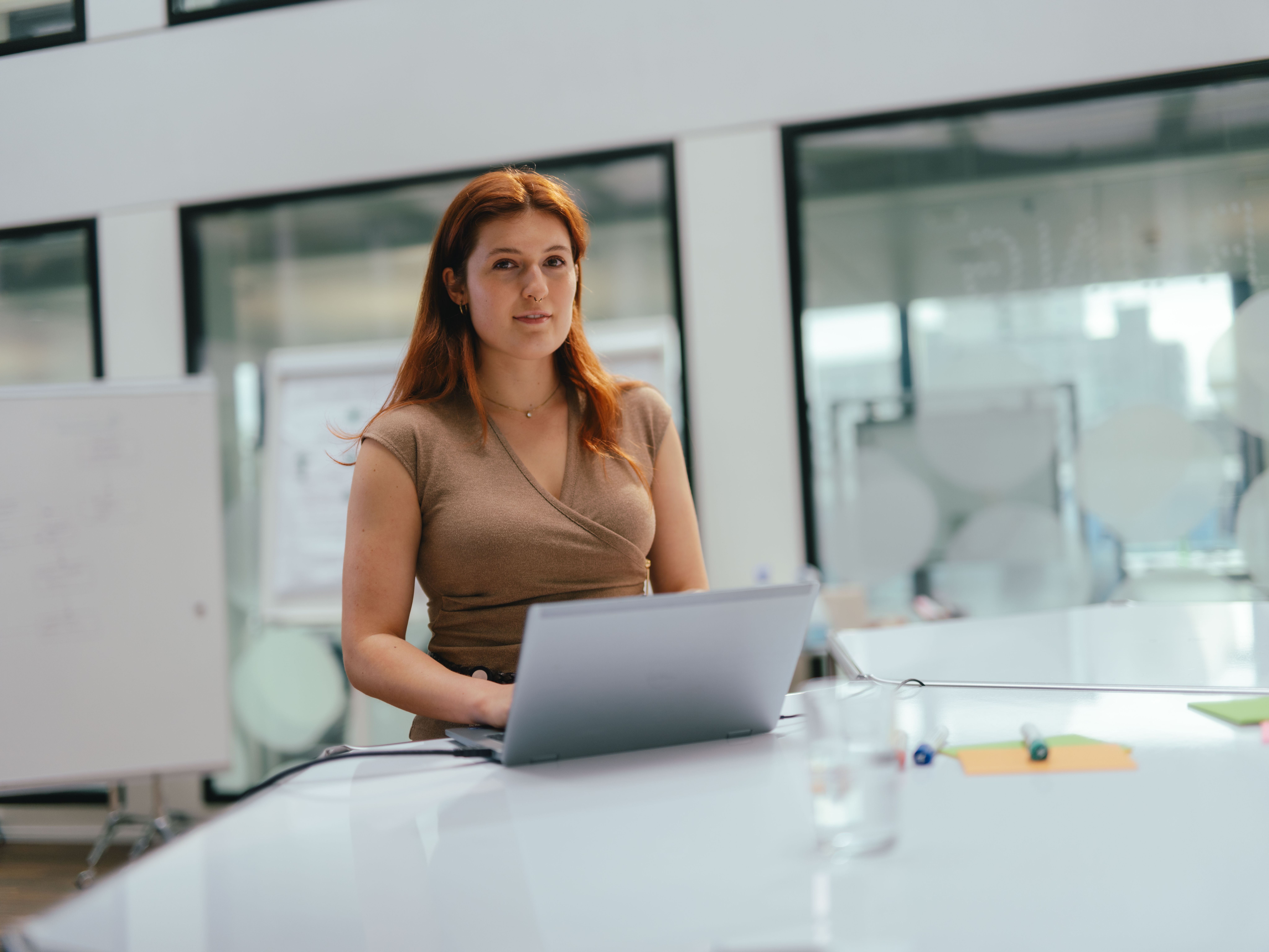 Eine Person steht an einem Tisch in einem modernen Bürobereich und arbeitet an einem aufgeklappten Laptop. Auf dem Tisch liegen ein Stift und eine kleine Wasserflasche. Im Hintergrund sind grosse Glaswände und weitere Büroräume sichtbar, sowie eine beschriftbare Tafel mit Notizen.