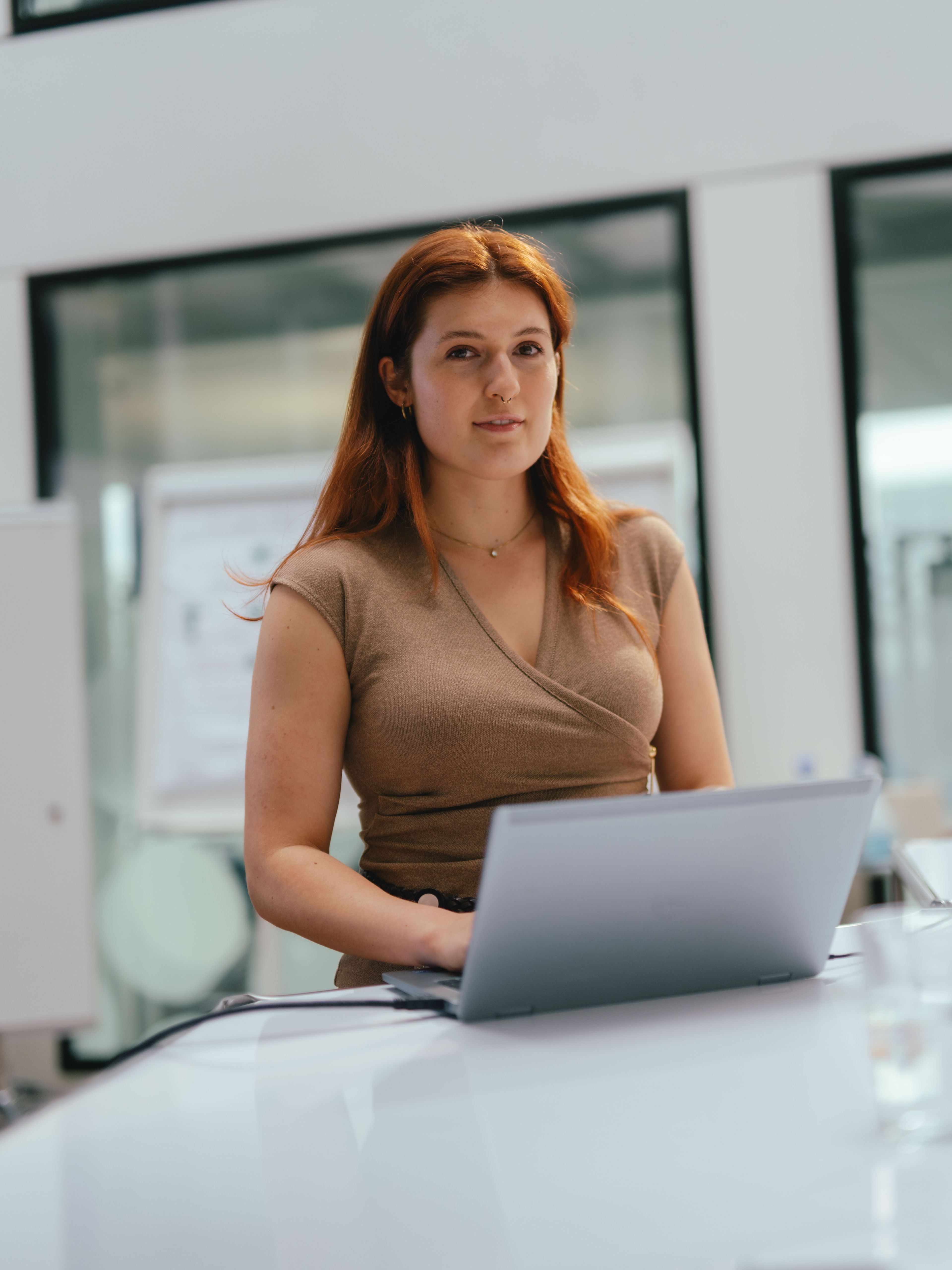 A person is seated at a large white conference table in a modern office, with an open laptop in front of them. In the background, there are glass walls, a whiteboard, and additional office elements. Pens and a water bottle are placed on the table.