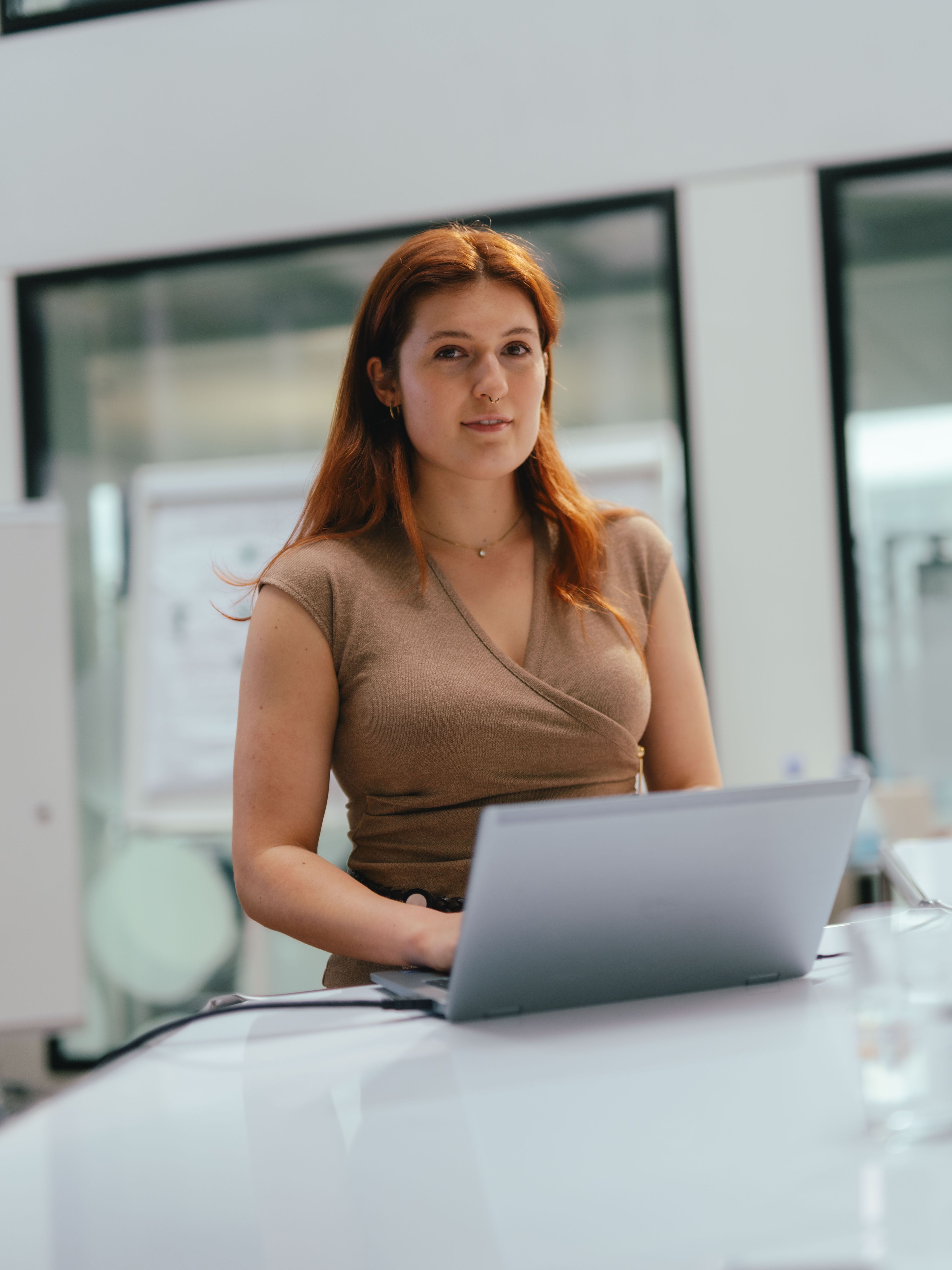 Eine Person steht an einem Tisch in einem modernen Bürobereich und arbeitet an einem aufgeklappten Laptop. Auf dem Tisch liegen ein Stift und eine kleine Wasserflasche. Im Hintergrund sind grosse Glaswände und weitere Büroräume sichtbar, sowie eine beschriftbare Tafel mit Notizen.