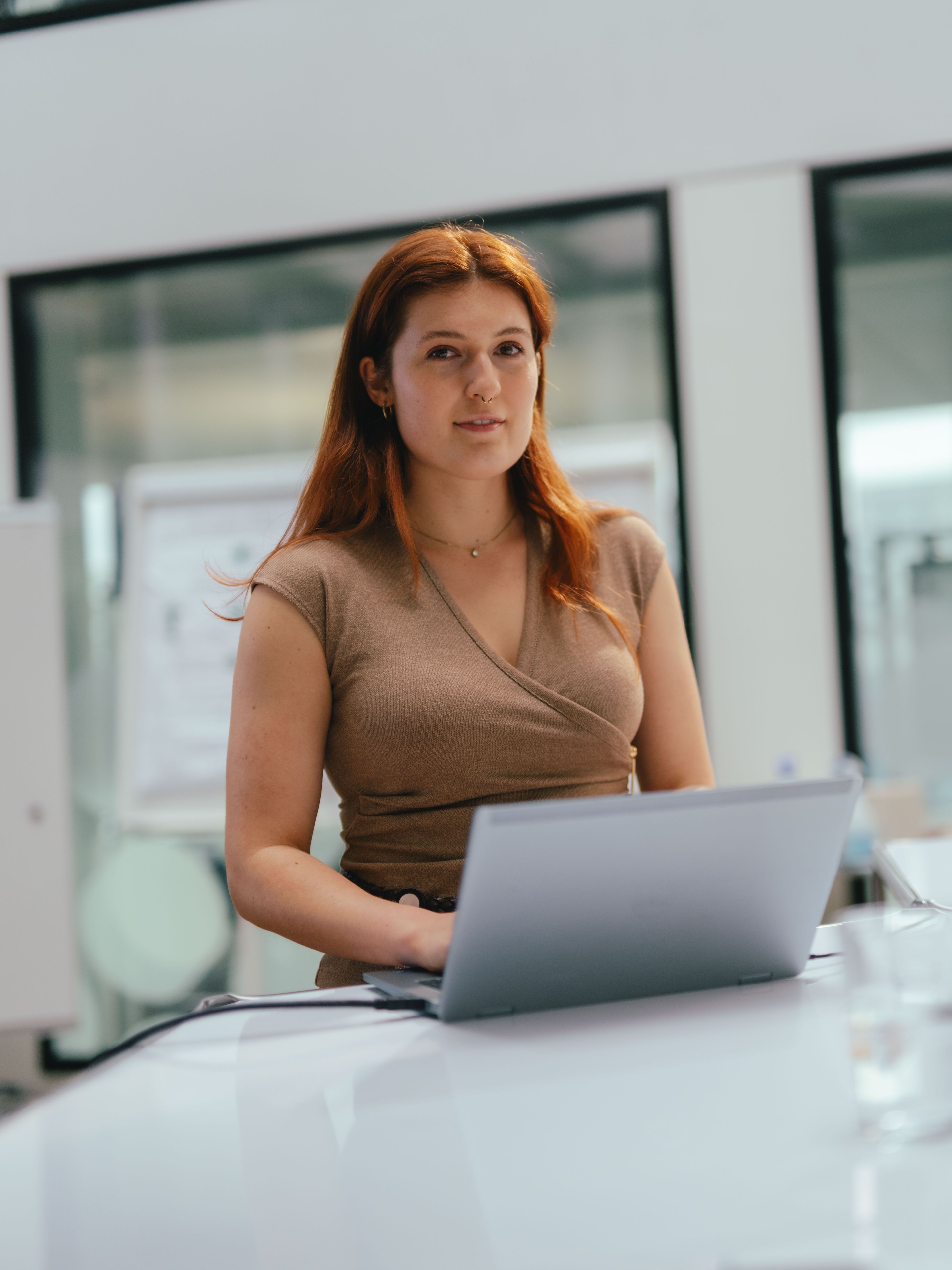 Une personne est assise à une grande table blanche dans un bureau moderne, avec un ordinateur portable ouvert devant elle. À l’arrière‑plan se trouvent des parois vitrées, un tableau blanc et d’autres éléments de bureau. Des stylos et une bouteille d’eau sont posés sur la table.