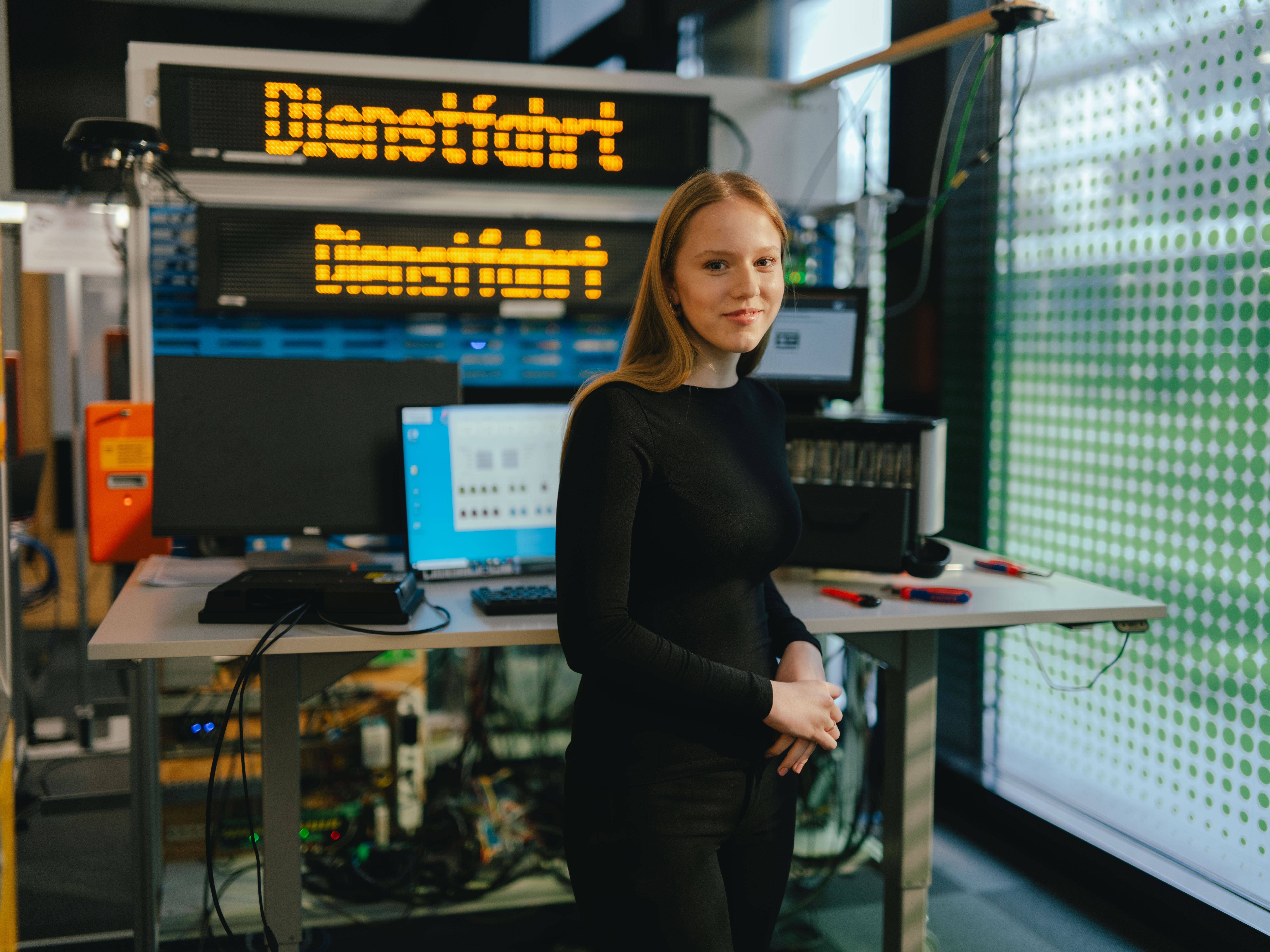 A person stands at a table in a technical room or lab. On the table are two open laptops and several electronic devices. In the background, a large digital display shows the yellow text “Dienstfahrt.” The room contains various technical equipment and cables.