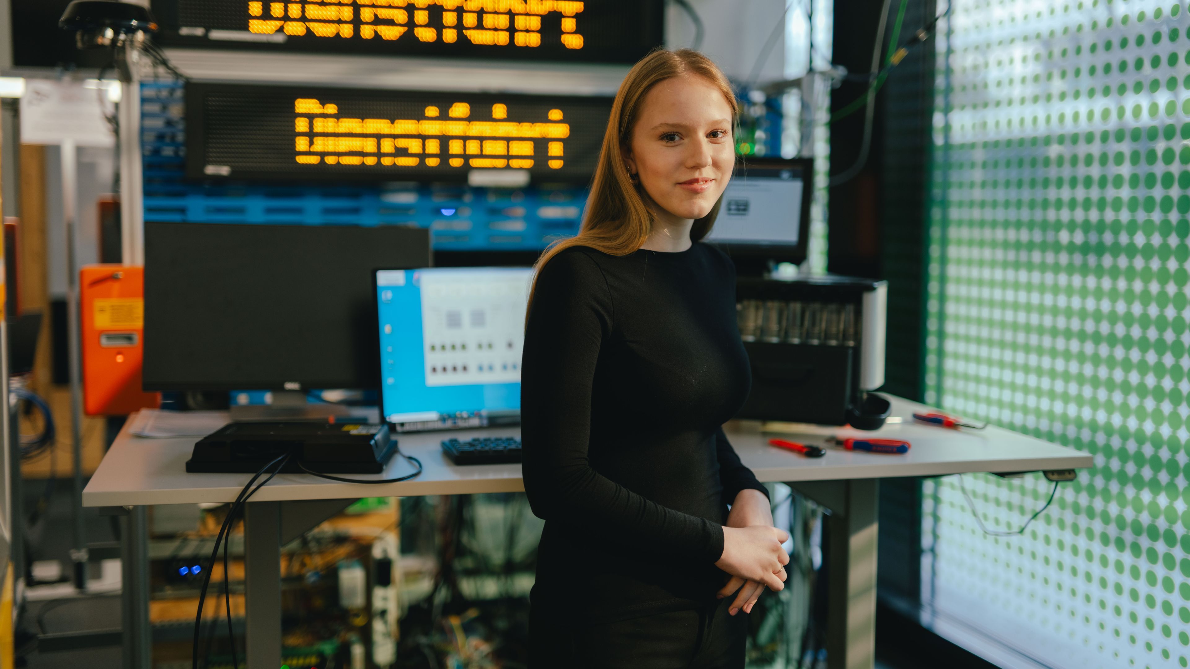 A person stands at a table in a technical room or lab. On the table are two open laptops and several electronic devices. In the background, a large digital display shows the yellow text “Dienstfahrt.” The room contains various technical equipment and cables.