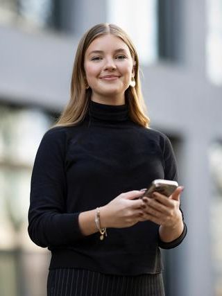 Eine Person steht im Freien vor einem modernen Gebäude und hält ein Smartphone in beiden Händen. Die Person trägt einen dunklen langen Pullover und ein Armband. Im Hintergrund ist eine gelbe Wand mit dem Post-Logo zu sehen.