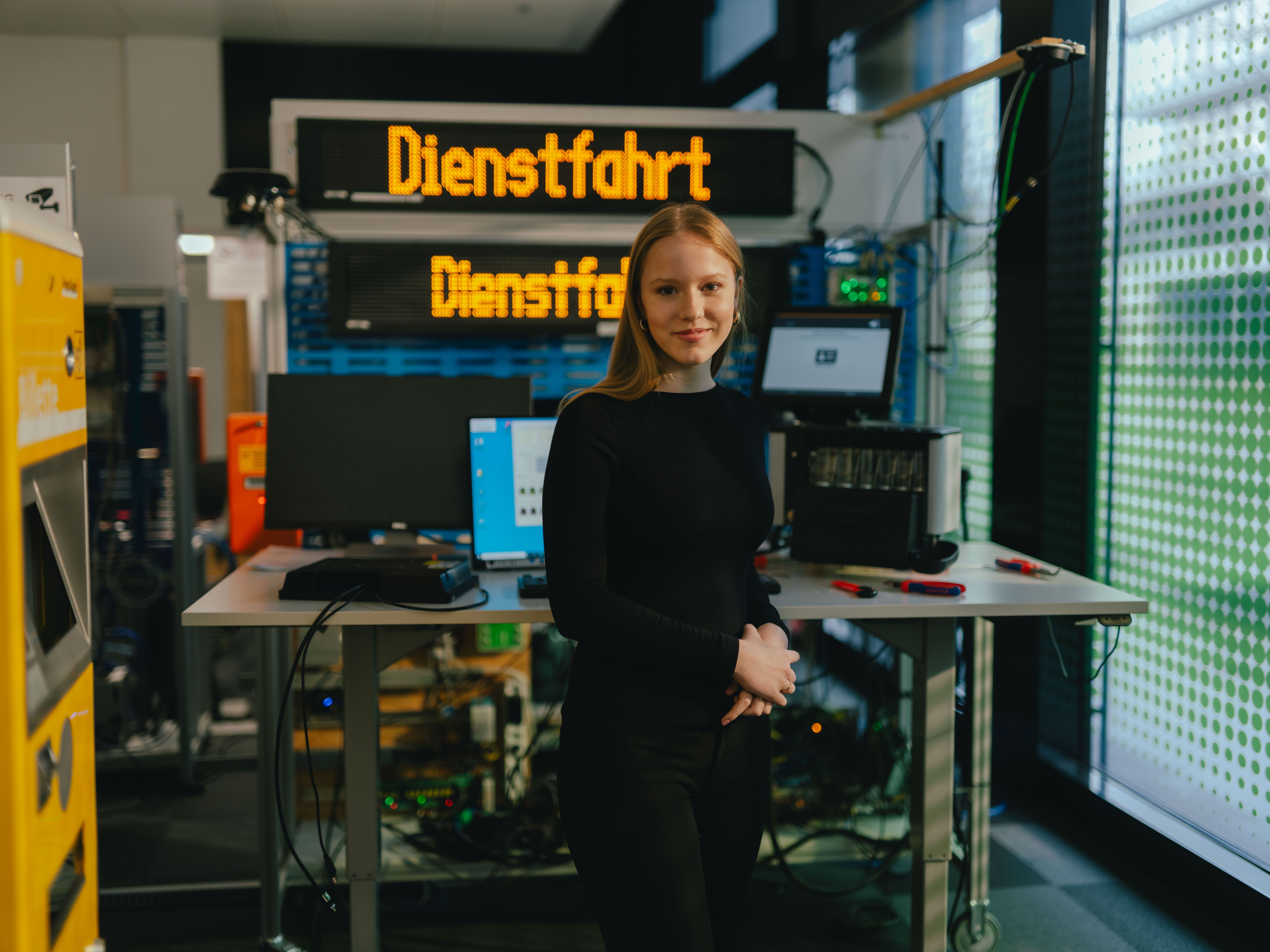 A person is standing in a technical workspace in front of a table with multiple monitors, computers, and electronic devices. Behind them is a large digital display board showing the yellow text “Dienstfahrt.” Additional technical equipment and a yellow device are visible on the left. The setting resembles a testing or workshop environment.