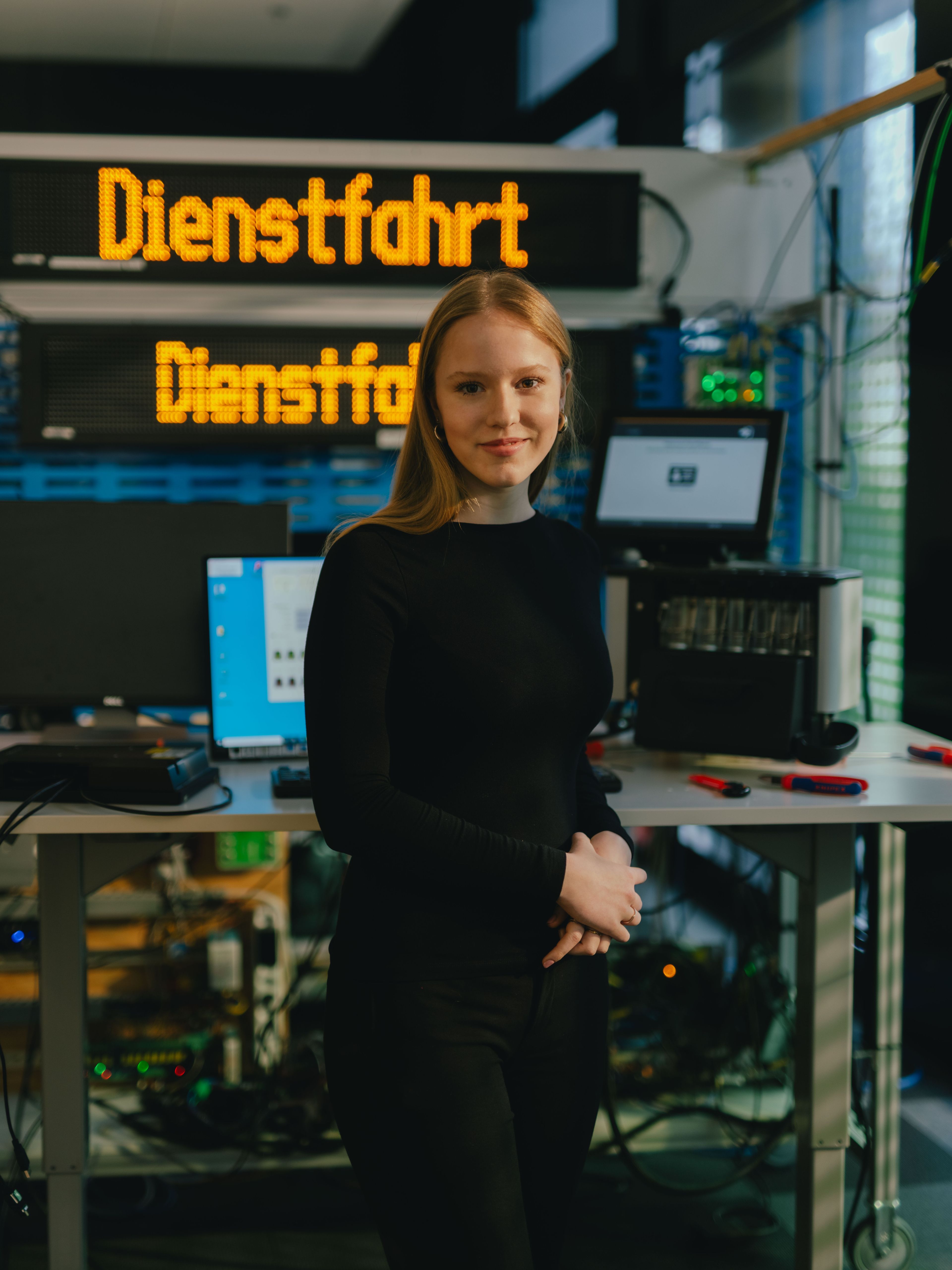 A person is standing in a technical workspace in front of a table with multiple monitors, computers, and electronic devices. Behind them is a large digital display board showing the yellow text “Dienstfahrt.” Additional technical equipment and a yellow device are visible on the left. The setting resembles a testing or workshop environment.