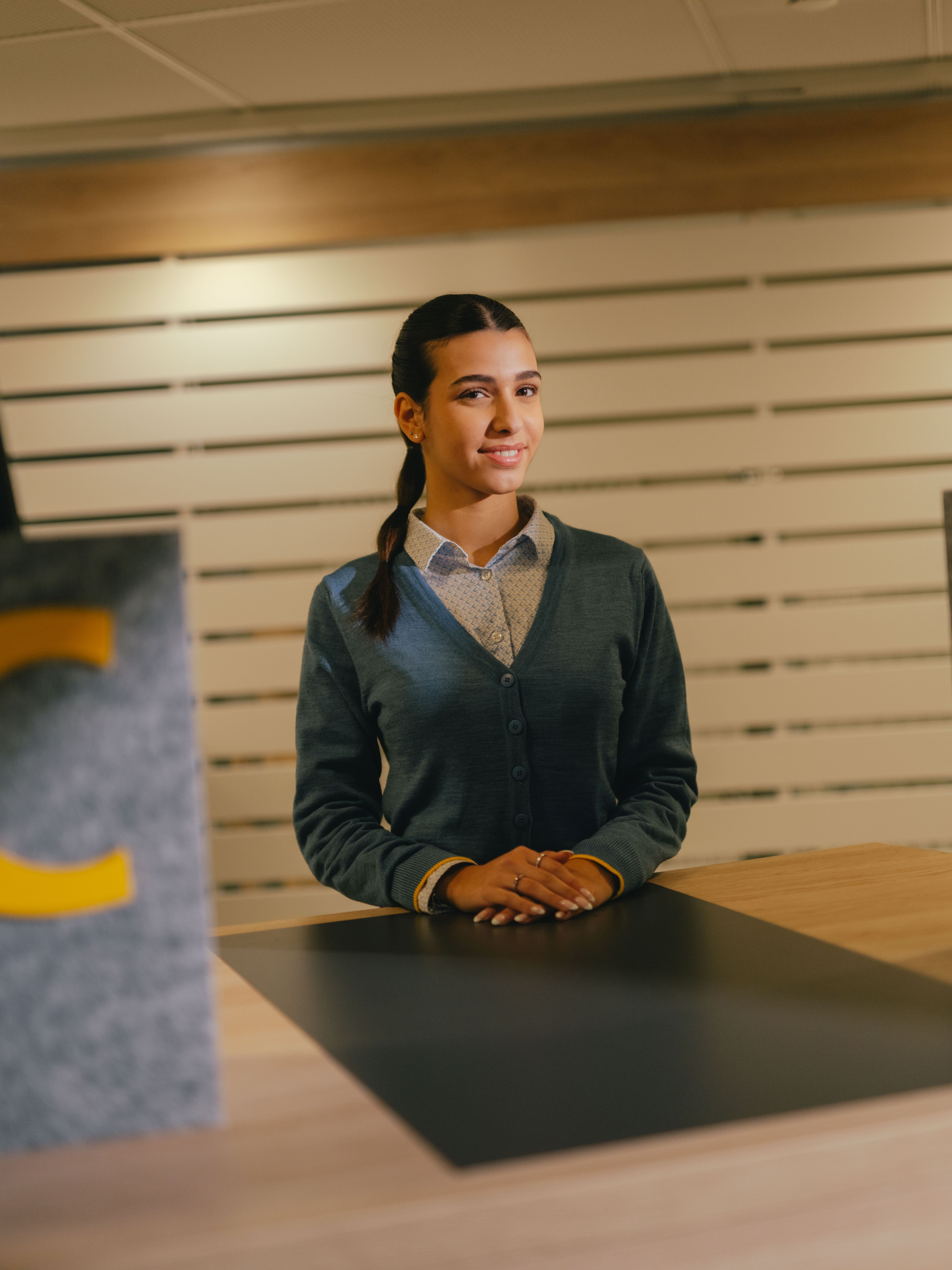 Eine Person steht hinter einem Tresen in einem Empfangs‑ oder Servicebereich. Vor der Person befindet sich eine dunkle Arbeitsfläche, eingerahmt von zwei grauen, strukturierten Elementen, von denen eines einen grossen gelben Buchstaben „C“ trägt. Der Hintergrund besteht aus horizontal angeordneten hellen Wandlamellen. Die Person trägt ein helles Hemd und eine dunkle Strickjacke und hat die Hände vor dem Körper abgelegt.