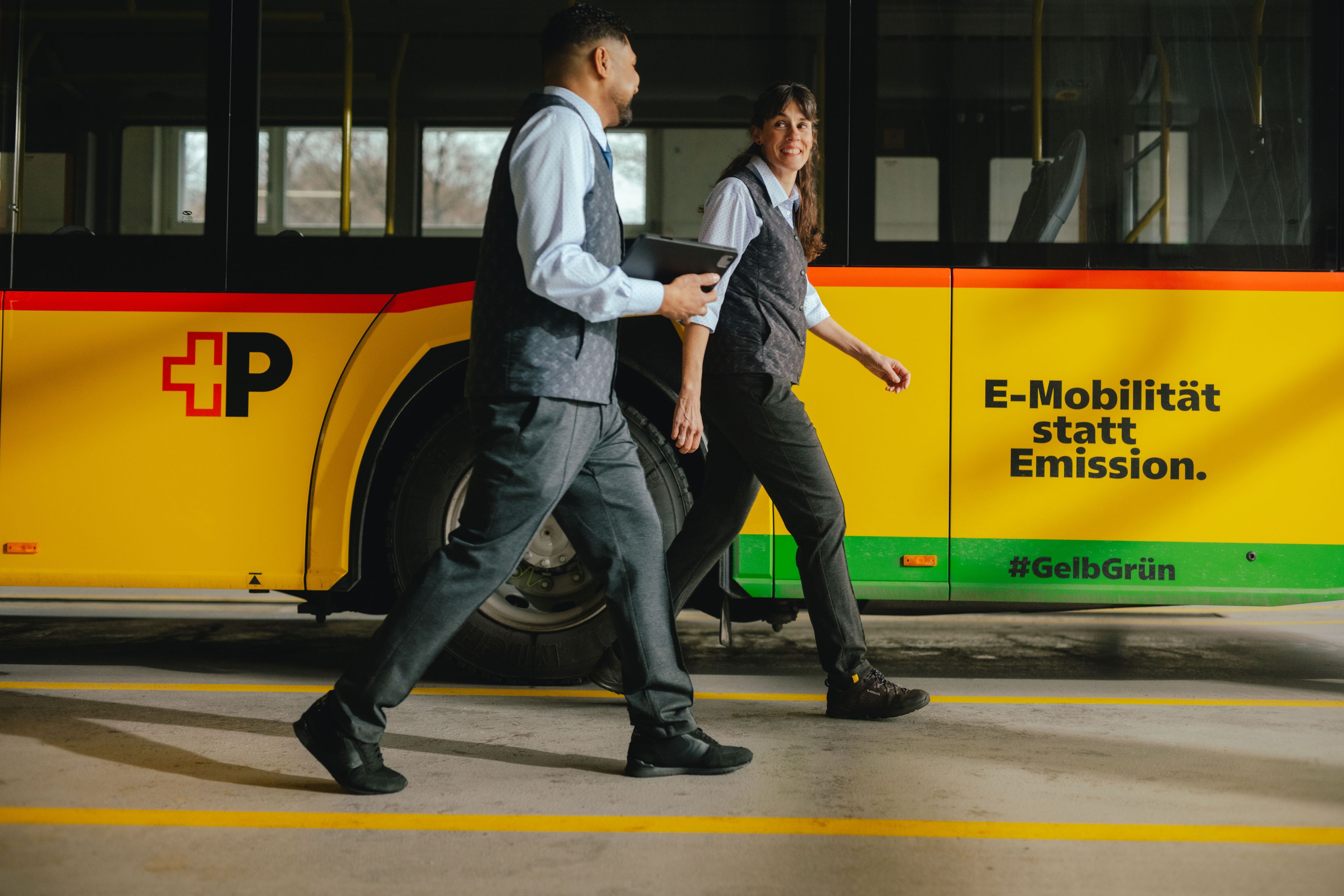Two people walk side by side in a hall next to a yellow and black bus. One of the individuals is holding a tablet. The side of the bus displays the text “E‑Mobilität statt Emission.” along with hashtags and coloured stripes. The setting appears to be a depot or maintenance area.