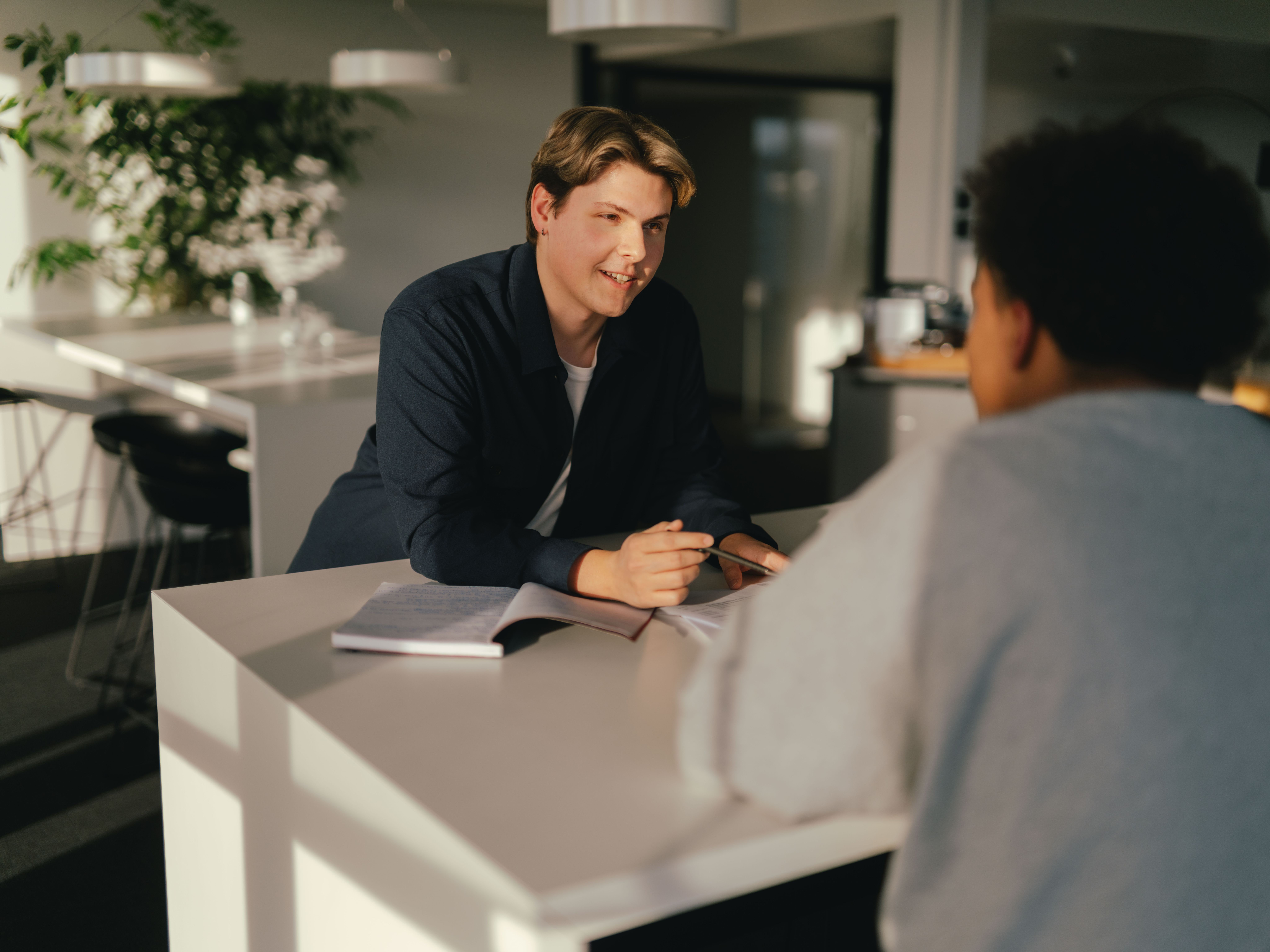 Zwei Personen sitzen sich an einem Tisch in einem modernen Innenraum gegenüber. Eine der Personen hält einen Stift über einem geöffneten Notizbuch, als ob etwas erklärt oder besprochen wird. Auf dem Tisch liegen weitere Unterlagen, und im Hintergrund sind Pflanzen, Möbel und grosse Fenster sichtbar. Das Licht fällt seitlich in den Raum und erzeugt weiche Schatten.