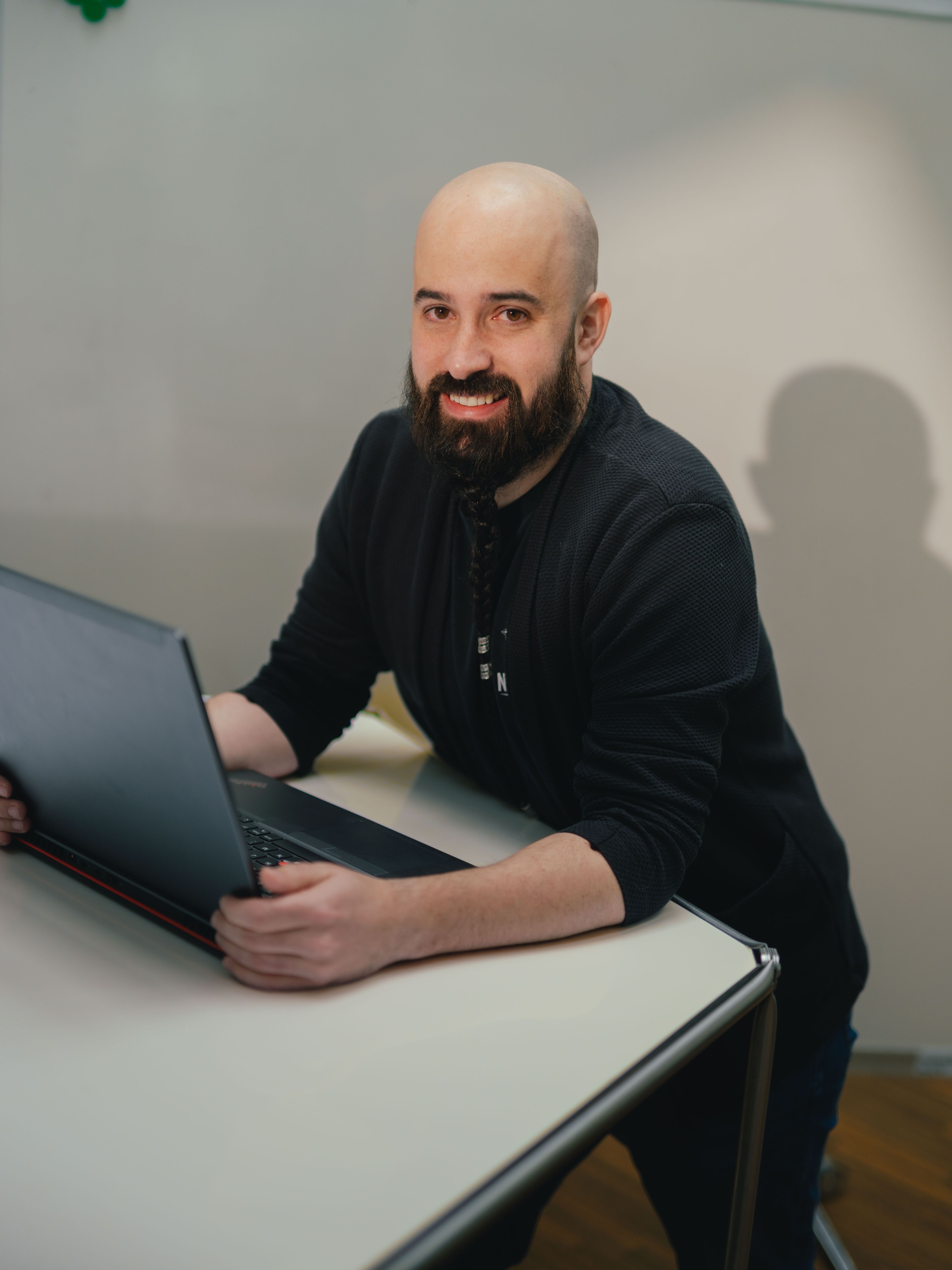 A person is seated at a table working on an open laptop. The person is wearing dark clothing. In the background, a glass wall with vertical structural elements and part of another room are visible. The table is light‑coloured with a smooth surface.