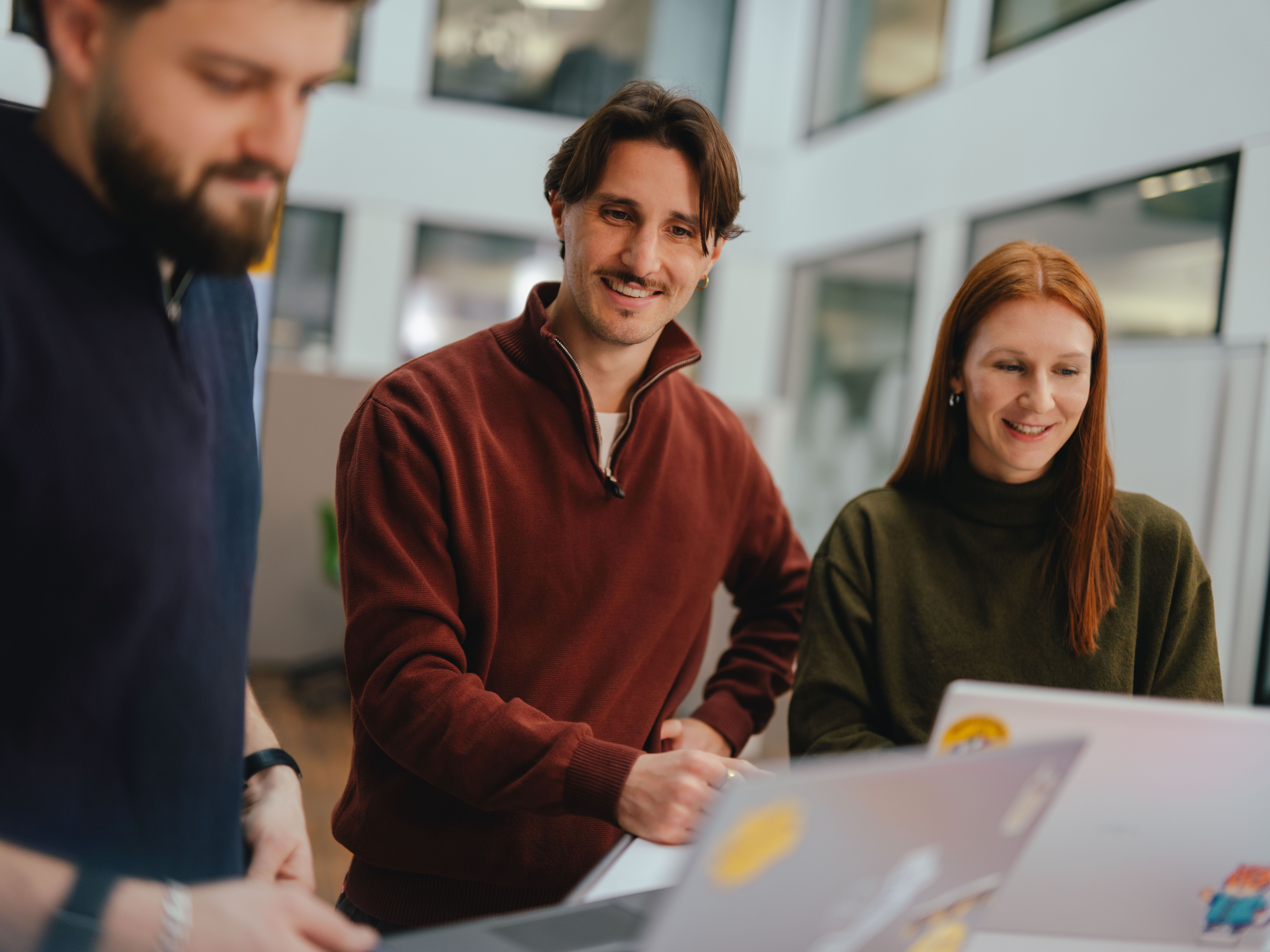 Three people are gathered around a workspace in a modern office. Open laptops are placed in front of them, and they appear to be collaborating or discussing something. In the background, there are glass partitions and office furnishings.