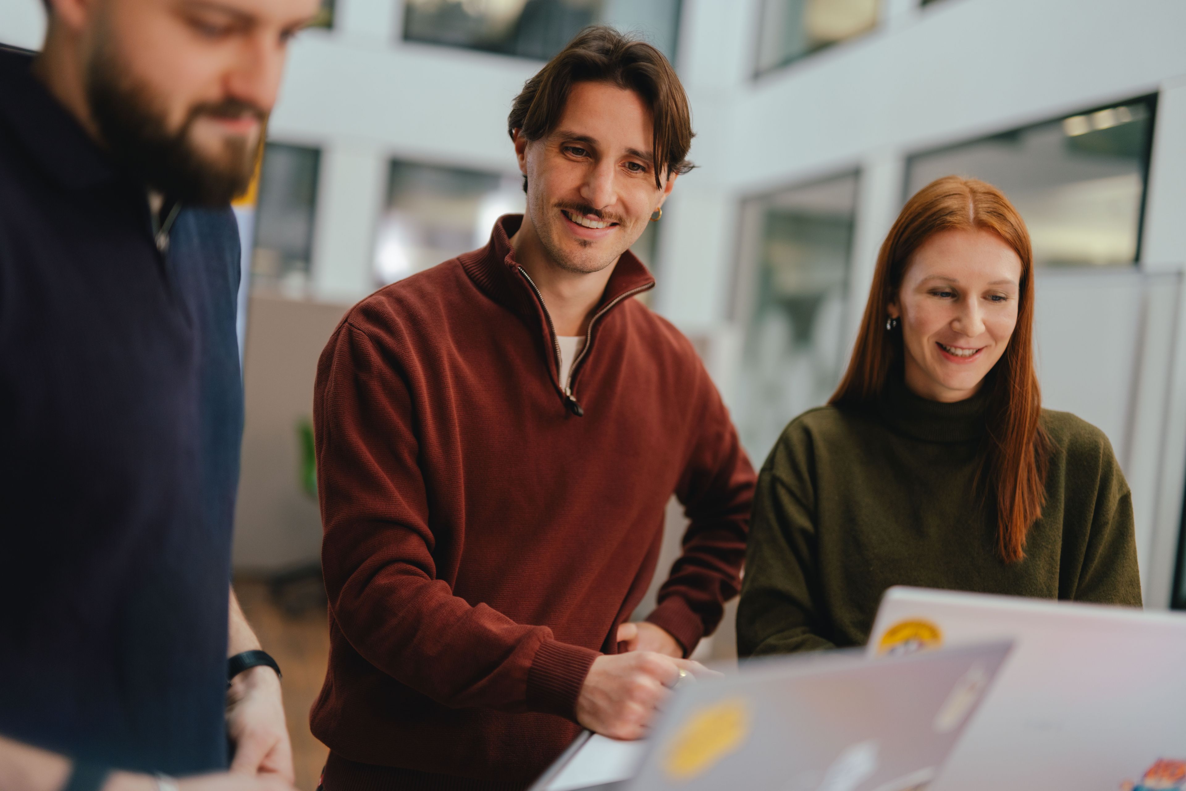 Three people are gathered around a workspace in a modern office. Open laptops are placed in front of them, and they appear to be collaborating or discussing something. In the background, there are glass partitions and office furnishings.