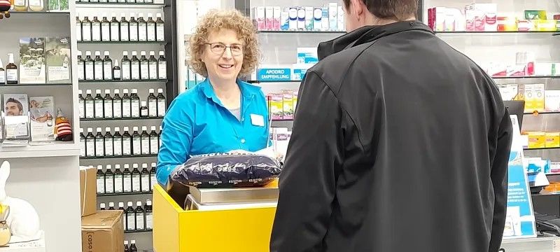 Service postal dans une pharmacie : une personne se tient au guichet postal jaune et dépose un colis. À l’arrière-plan, des étagères avec des médicaments, des flacons et des produits de santé.
