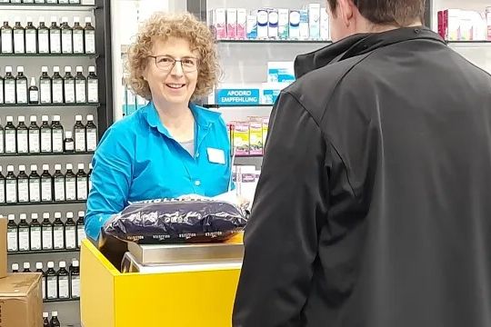 Service postal dans une pharmacie : une personne se tient au guichet postal jaune et dépose un colis. À l’arrière-plan, des étagères avec des médicaments, des flacons et des produits de santé.