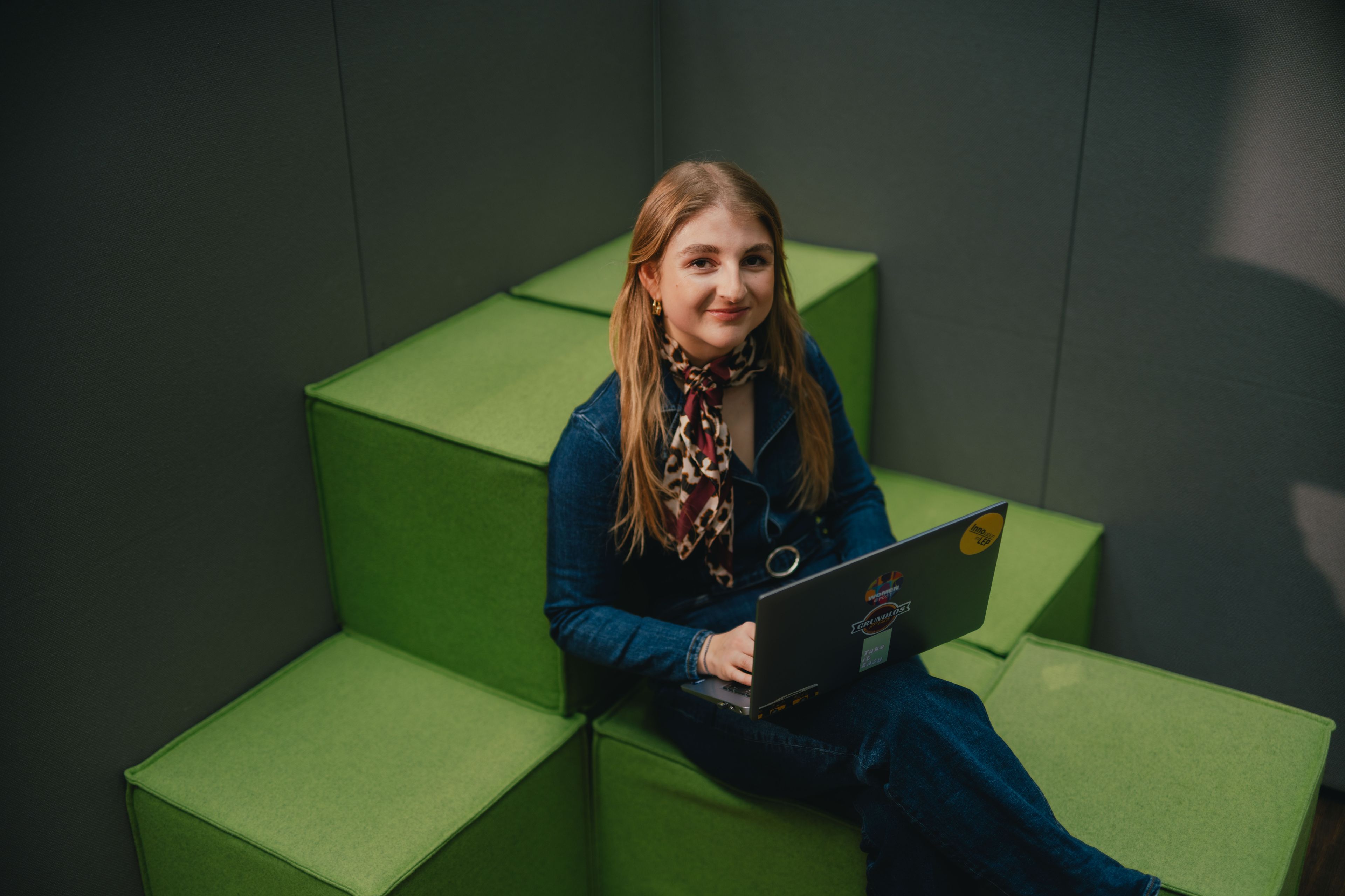 Eine Person sitzt auf grünen, stufenförmig angeordneten Polsterelementen und arbeitet mit einem aufgeklappten Laptop auf dem Schoss. Die Person trägt dunkle Kleidung und ein gemustertes Tuch um den Hals. Der Raum wirkt gedämpft beleuchtet, und die Wände um die Sitzfläche sind dunkel gehalten.