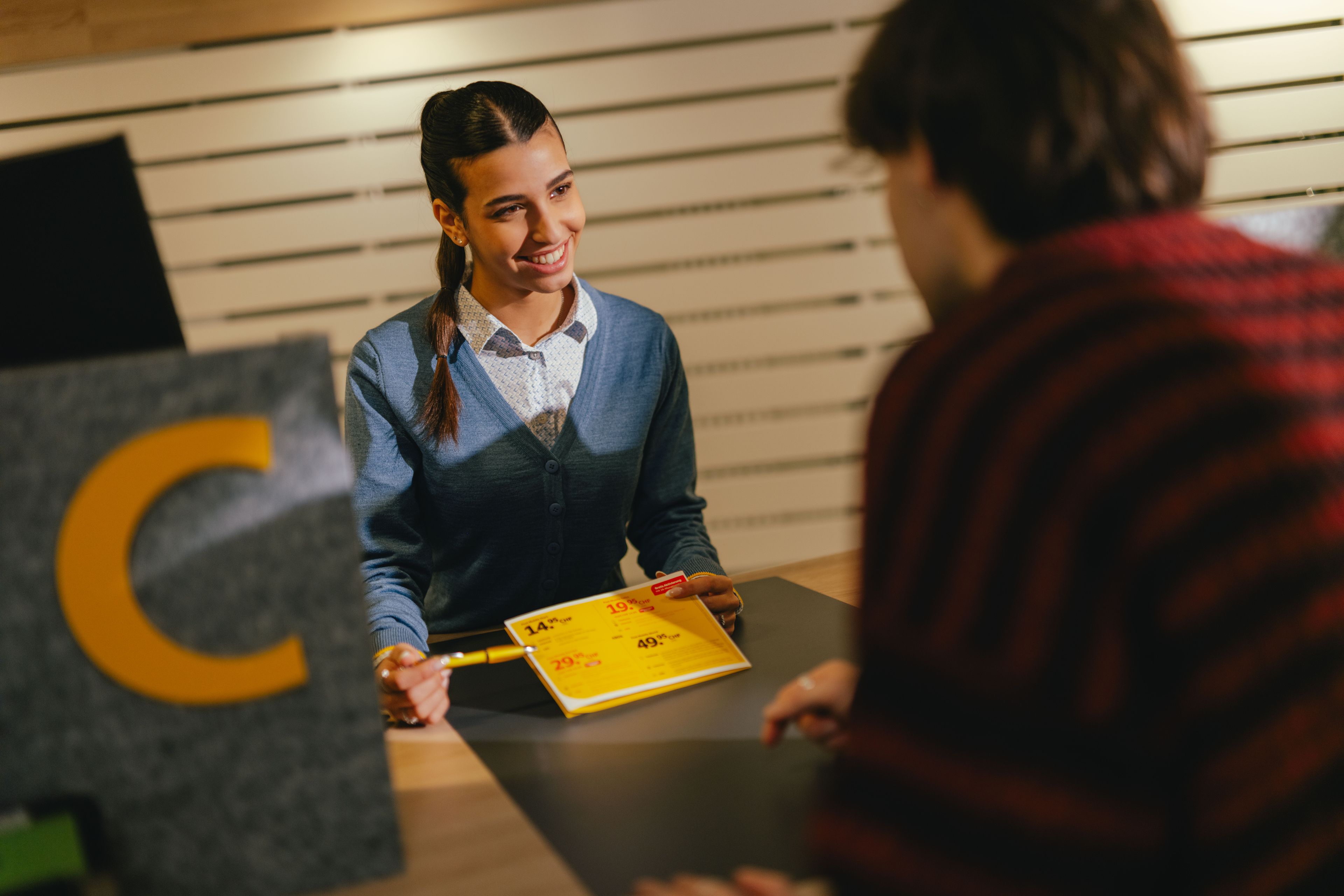 Eine Person sitzt an einem Schalter und reicht ein gelbes Formular über den Tresen an eine andere Person. Der Tresenbereich ist mit einem grauen Element markiert, das einen grossen gelben Buchstaben „C“ zeigt. Die Umgebung wirkt wie ein Empfangs‑ oder Servicebereich mit hellen Holzlamellen im Hintergrund.