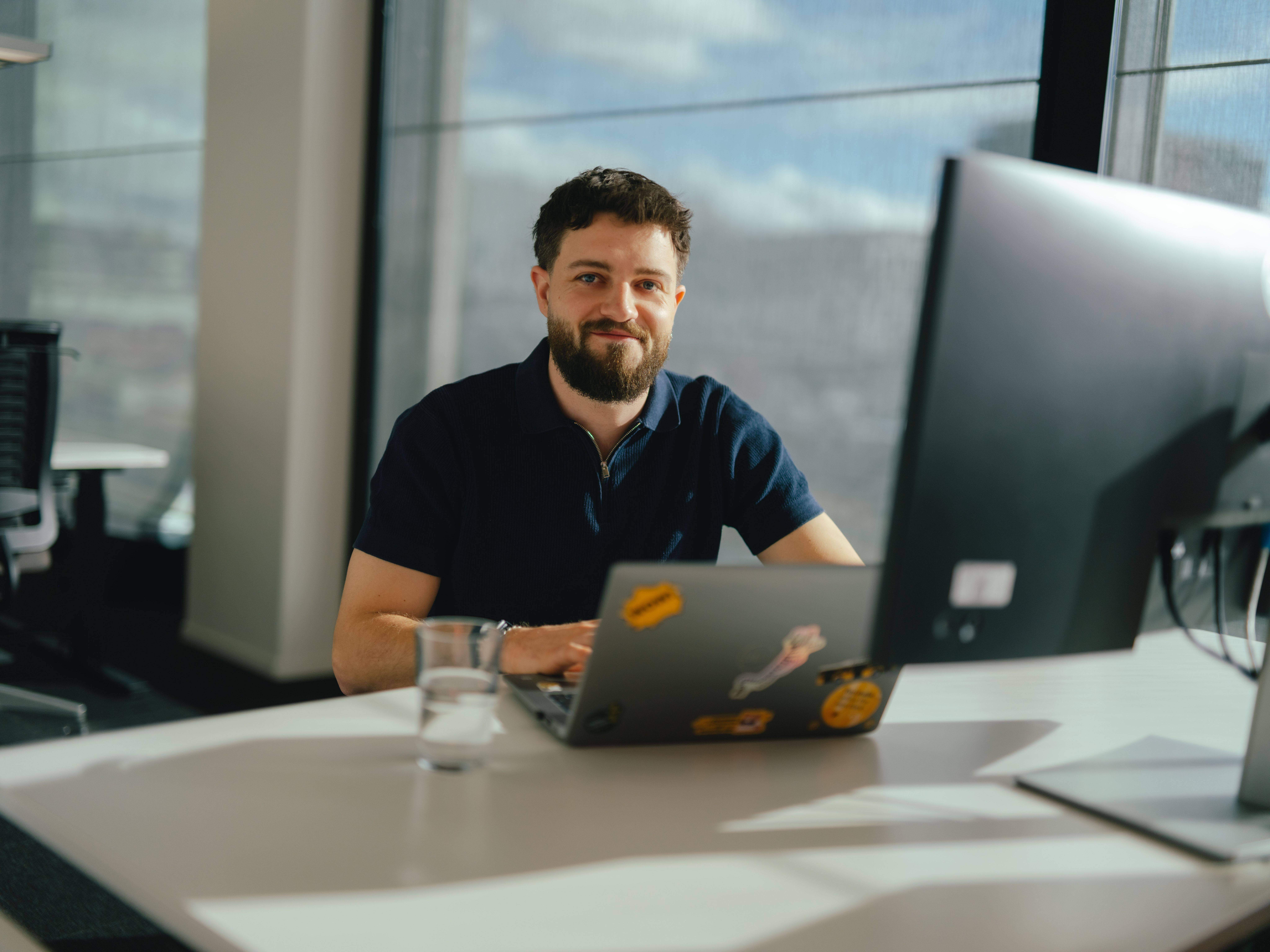 Eine Person sitzt an einem Schreibtisch und arbeitet an einem Laptop, der mit mehreren Aufklebern dekoriert ist. Vor der Person stehen ein grosses Computerdisplay und ein Wasserglas. Durch die grossen Fenster fällt helles Tageslicht in den Raum, und im Hintergrund sind weitere Büroarbeitsplätze sichtbar.