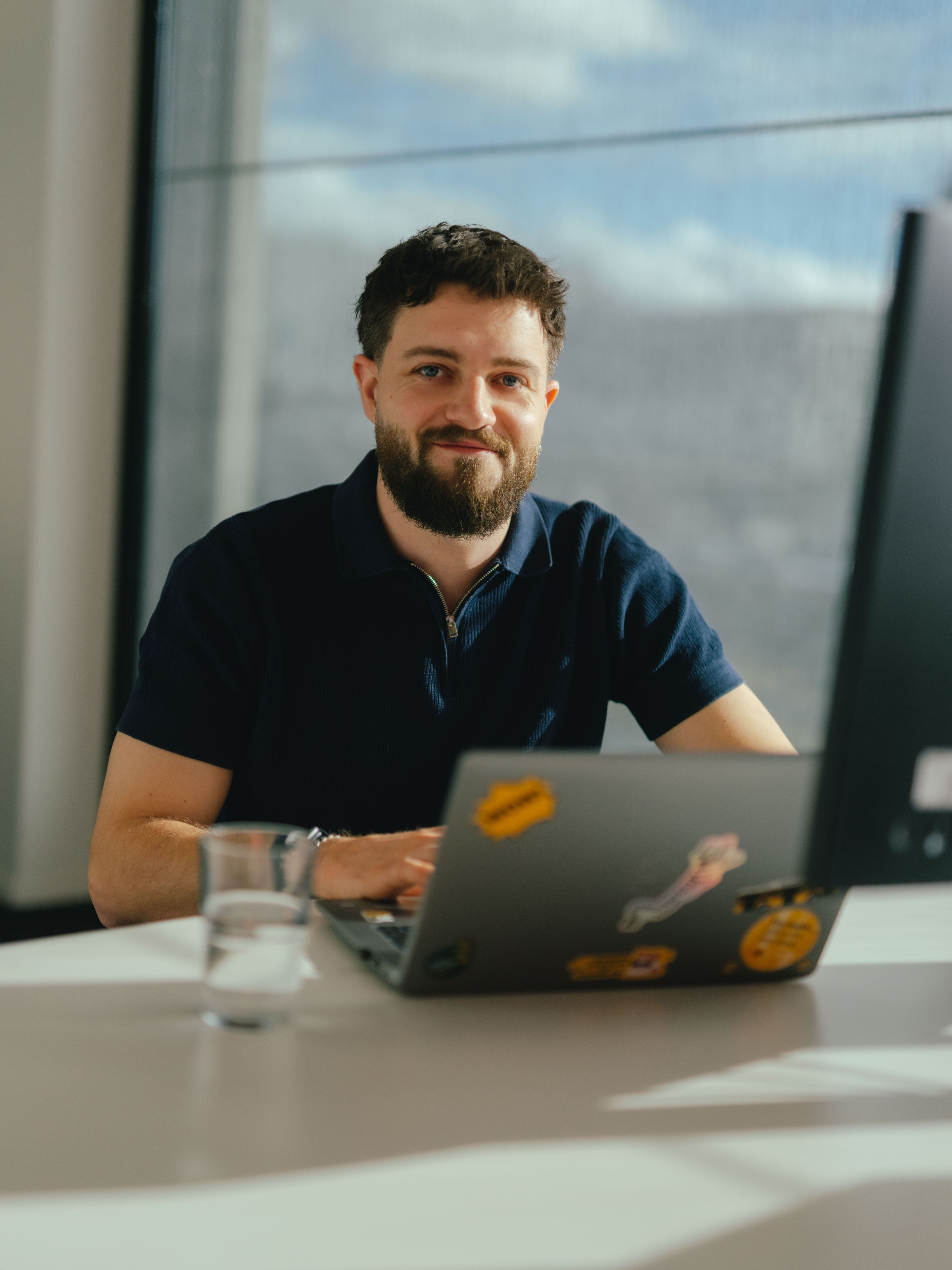 Eine Person sitzt an einem Schreibtisch und arbeitet an einem Laptop, der mit mehreren Aufklebern dekoriert ist. Vor der Person stehen ein grosses Computerdisplay und ein Wasserglas. Durch die grossen Fenster fällt helles Tageslicht in den Raum, und im Hintergrund sind weitere Büroarbeitsplätze sichtbar.