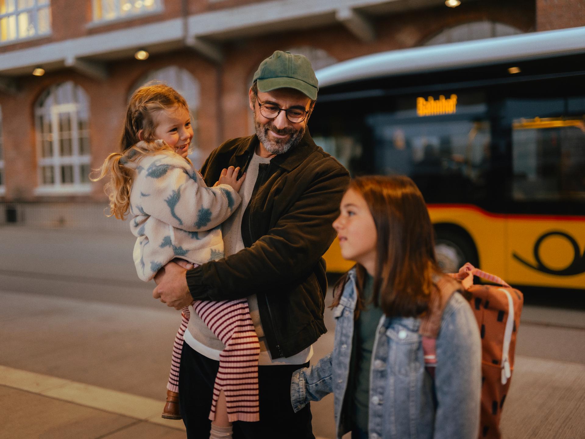 Drei Personen stehen an einer Haltestelle vor einem gelben Bus. Eine Person hält ein Kind im Arm, während eine weitere Person mit einem Rucksack daneben steht. Im Hintergrund ist ein Backsteingebäude mit beleuchteten Fenstern zu sehen.