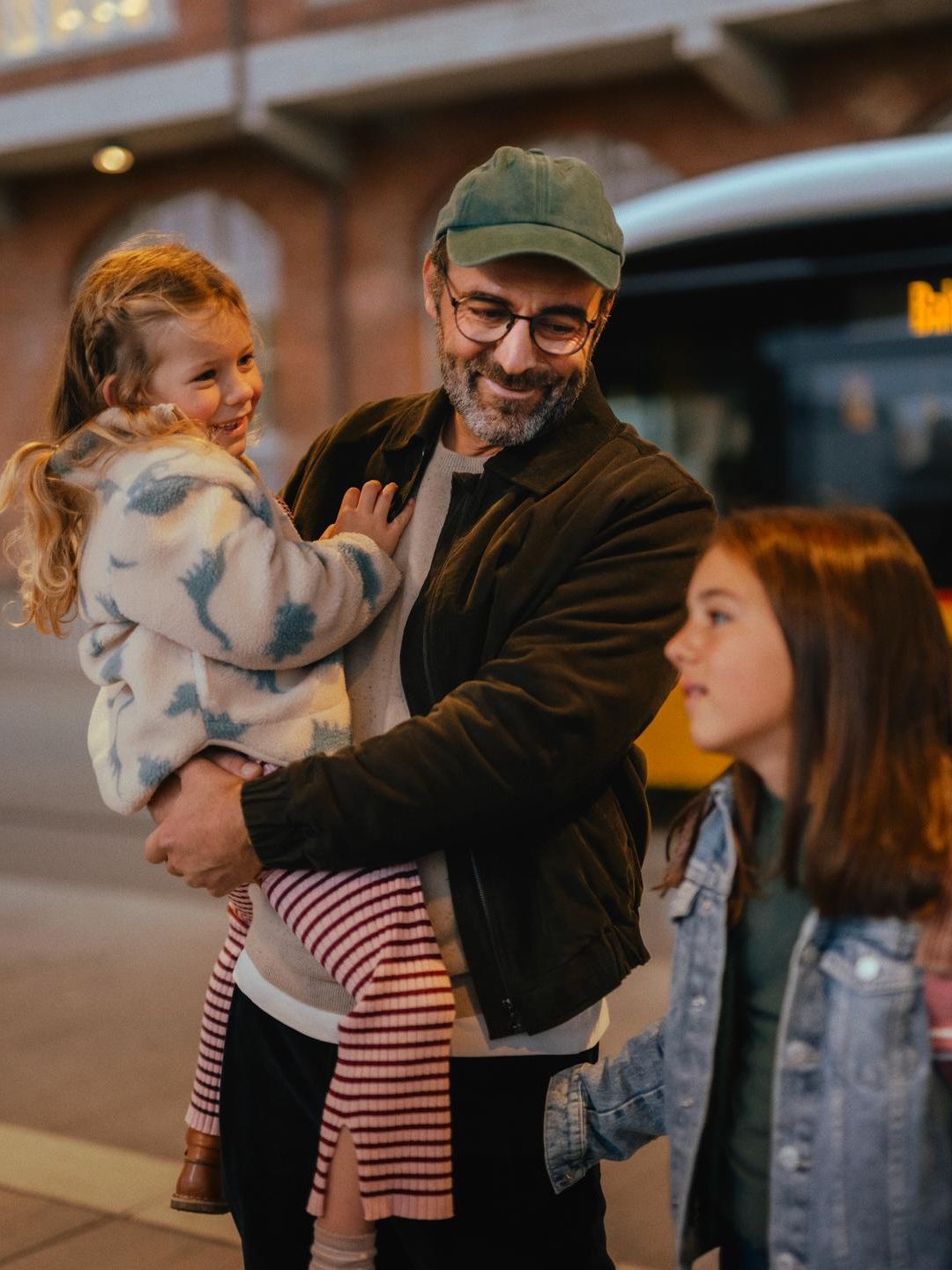 Three people are standing at a bus stop in front of a yellow bus. One person is holding a child, while another person with a backpack stands nearby. In the background, a brick building with illuminated windows is visible.