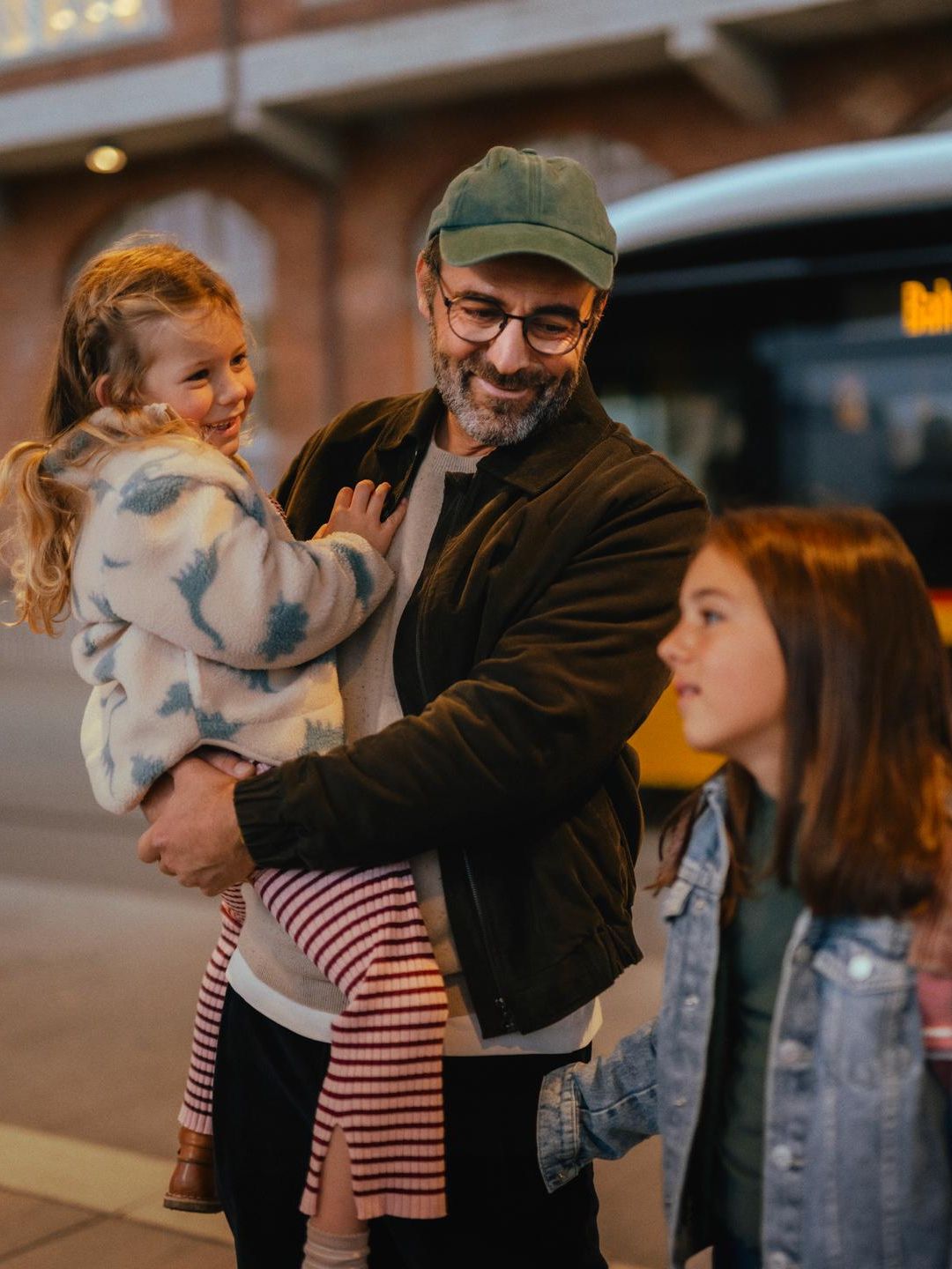 Trois personnes se tiennent à un arrêt de bus devant un bus jaune. Une personne porte un enfant dans les bras, tandis qu’une autre personne avec un sac à dos se tient à côté. En arrière‑plan, on voit un bâtiment en briques avec des fenêtres éclairées.