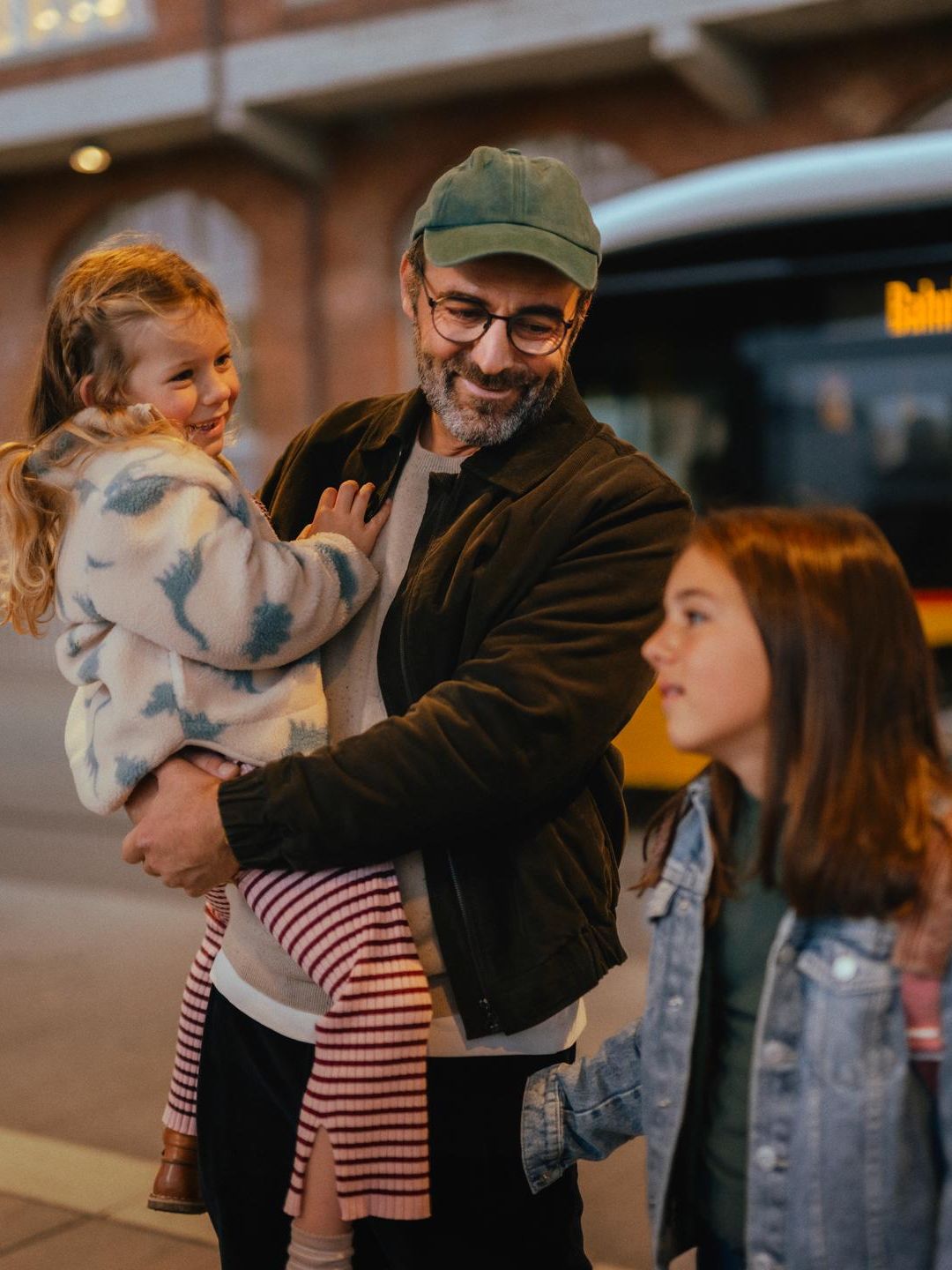 Drei Personen stehen an einer Haltestelle vor einem gelben Bus. Eine Person hält ein Kind im Arm, während eine weitere Person mit einem Rucksack daneben steht. Im Hintergrund ist ein Backsteingebäude mit beleuchteten Fenstern zu sehen.