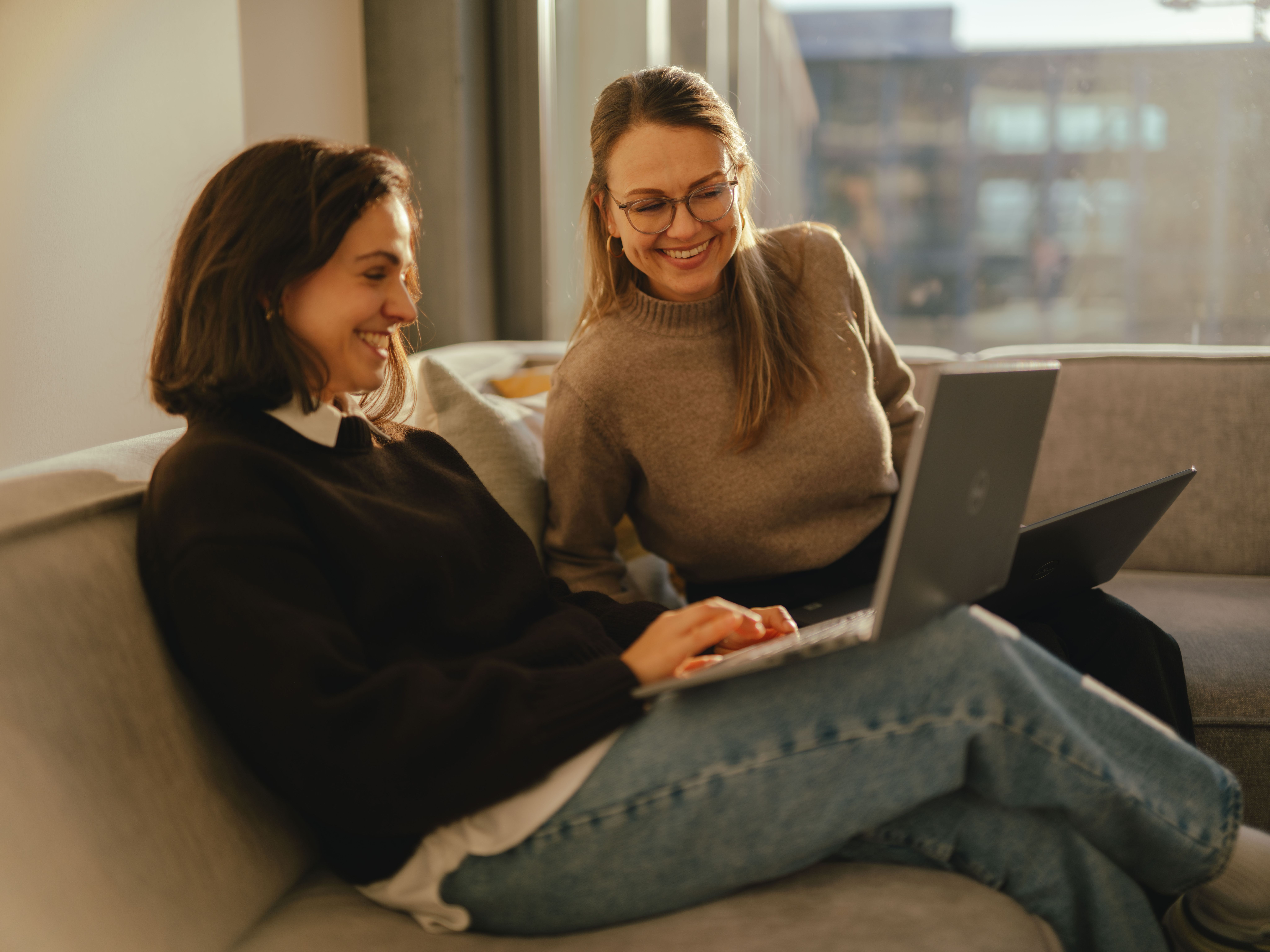 Deux personnes sont assises sur un canapé dans un espace intérieur moderne et travaillent chacune sur un ordinateur portable ouvert. Les deux sont assises côte à côte, l’un des ordinateurs reposant sur les jambes d’une personne. À l’arrière‑plan, de grandes fenêtres laissent apparaître des bâtiments urbains et une lumière chaude.