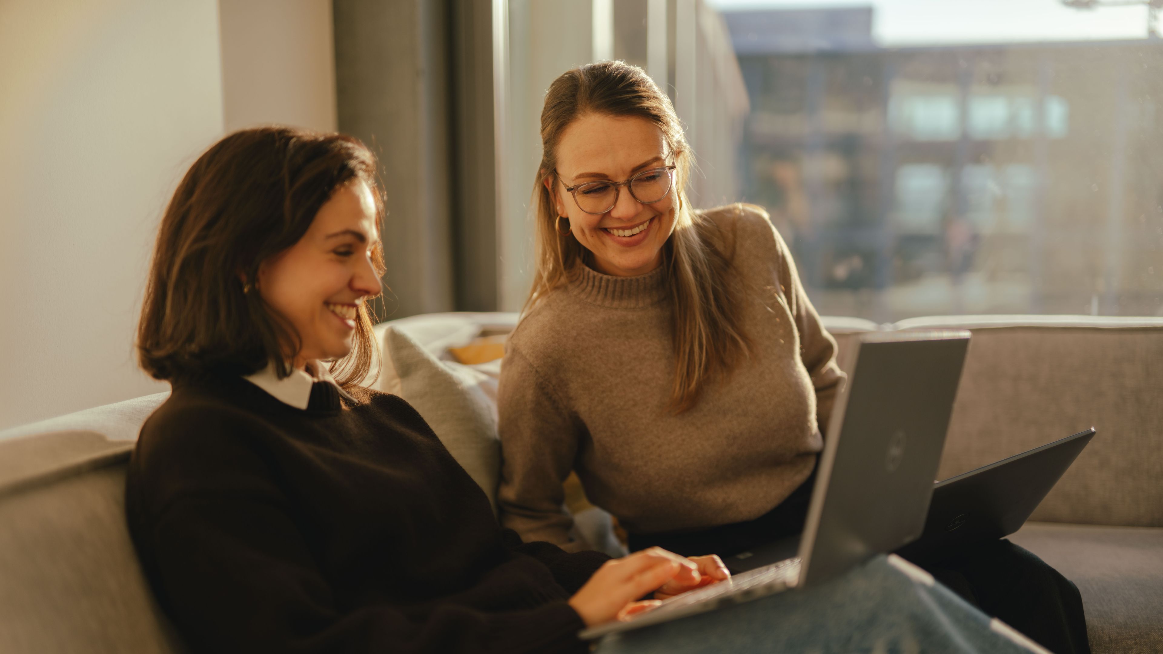 Zwei Personen sitzen auf einem Sofa in einem modernen Innenraum und arbeiten jeweils an aufgeklappten Laptops. Beide sitzen nahe beieinander, und einer der Laptops liegt auf den Oberschenkeln einer Person. Im Hintergrund sind grosse Fenster sichtbar, durch die eine städtische Gebäudestruktur und warmes Tageslicht in den Raum fallen.