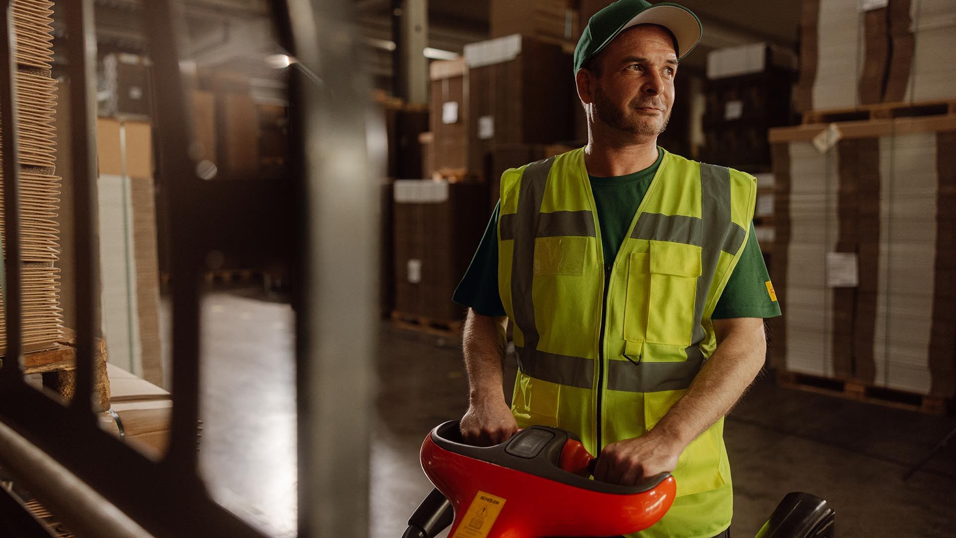 An employee in a high-visibility vest operates an electric pallet truck in a warehouse and transports pallets of goods between shelves.