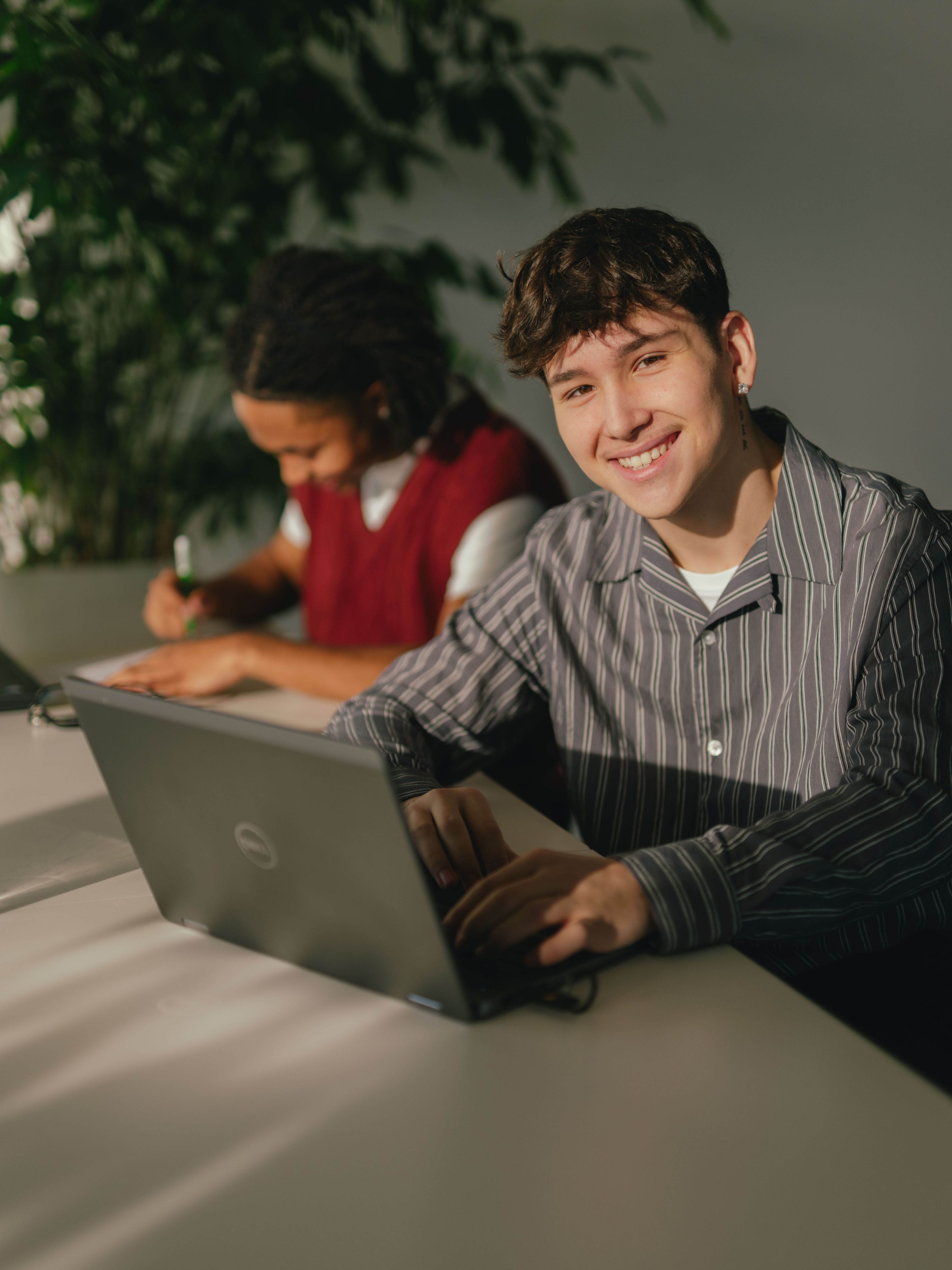 Deux personnes sont assises à une longue table et travaillent chacune sur un ordinateur portable. La personne au premier plan porte un haut rayé et utilise un ordinateur portable couleur argent. Un verre d’eau se trouve à côté. Derrière cette personne se trouve une autre personne assise à un ordinateur portable noir. À l’arrière-plan se trouve une grande plante verte, et la lumière du soleil pénètre de travers dans la pièce et crée des motifs d’ombre clairs sur la table et le mur.