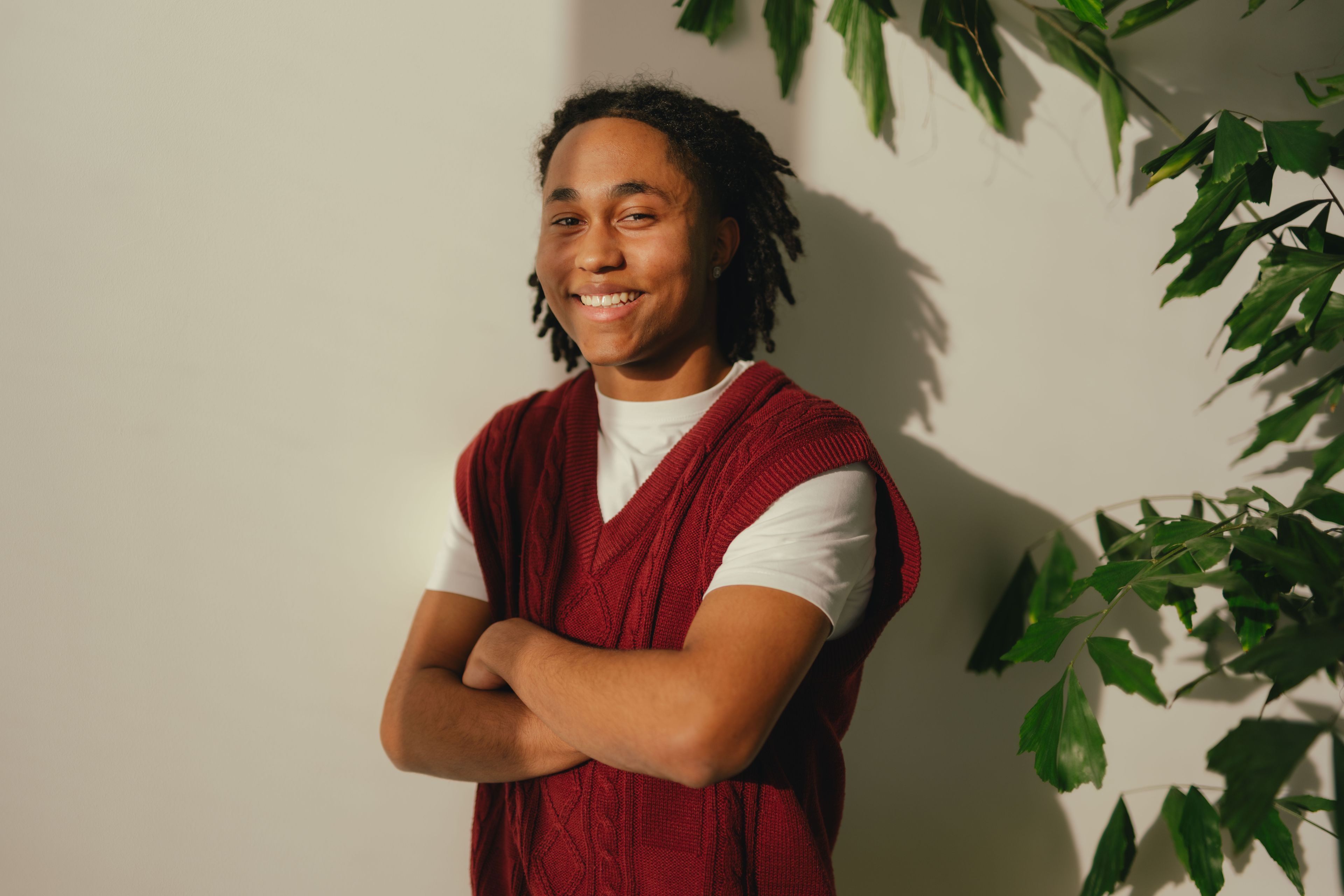 Une personne se tient devant un mur clair, entourée en partie par des ombres et des feuilles de plantes. La personne porte un T‑shirt blanc sous un pull sans manches en tricot rouge et garde les bras croisés.