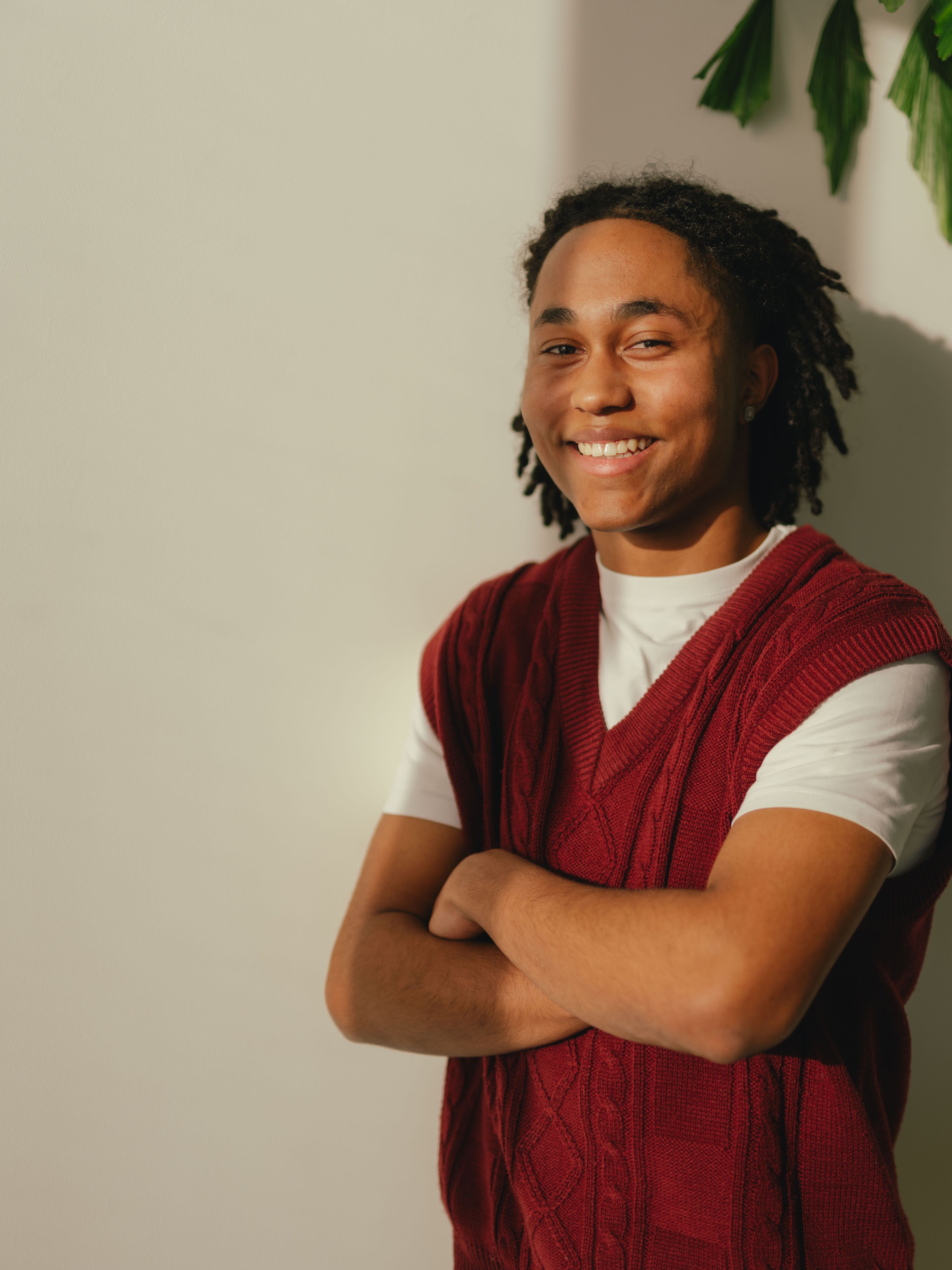 Eine Person steht vor einer hellen Wand und hat die Arme vor dem Körper verschränkt. Die Person trägt ein weisses T‑Shirt unter einem gestrickten ärmellosen Oberteil in dunklem Rot. Auf der rechten Bildseite ragen mehrere grüne Pflanzenblätter ins Bild.