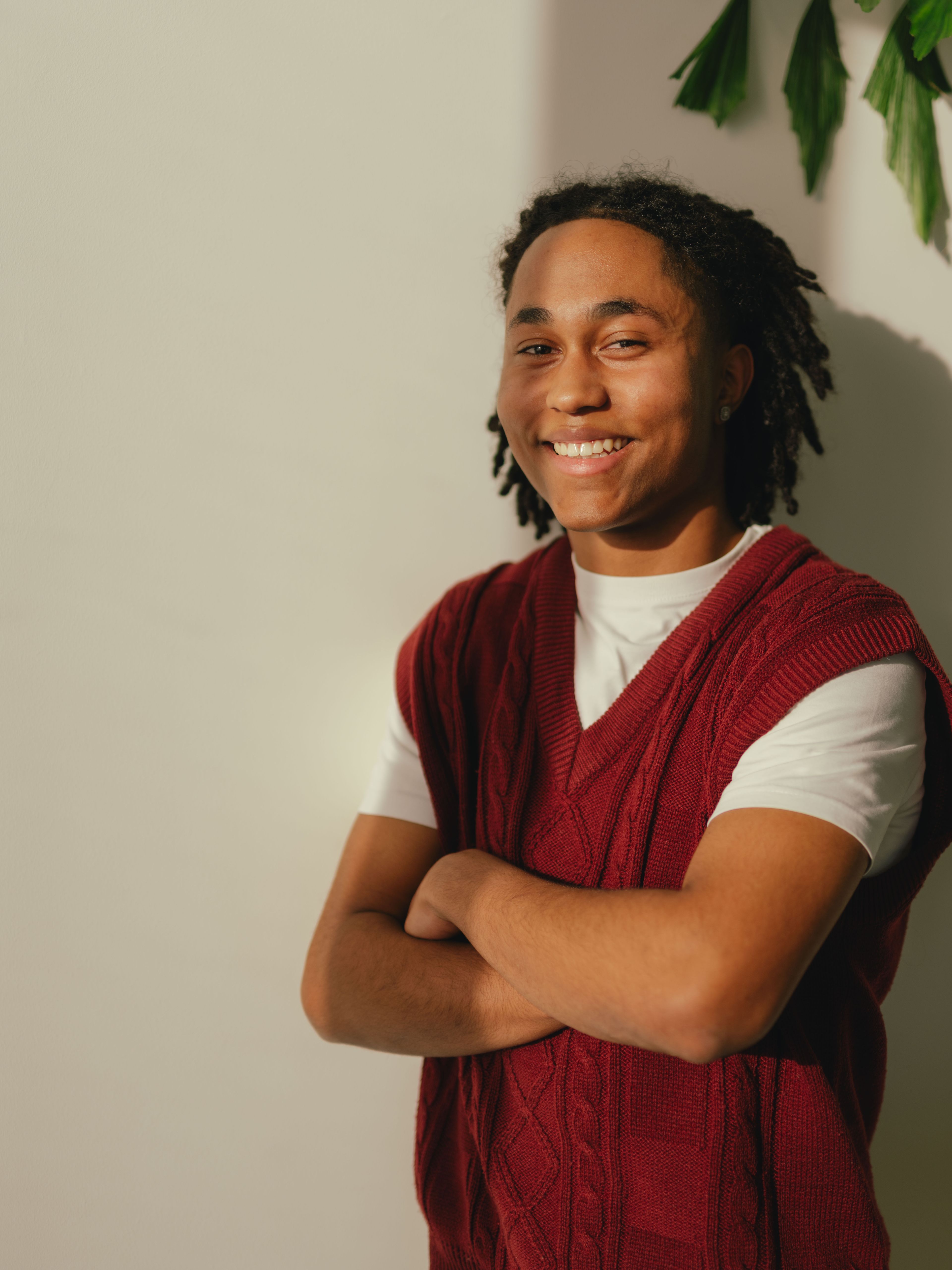Une personne se tient devant un mur clair et croise les bras devant son corps. La personne porte un t-shirt blanc sous un haut tricoté sans manches rouge foncé. Sur le côté droit de l’image, plusieurs feuilles de plantes vertes dépassent.
