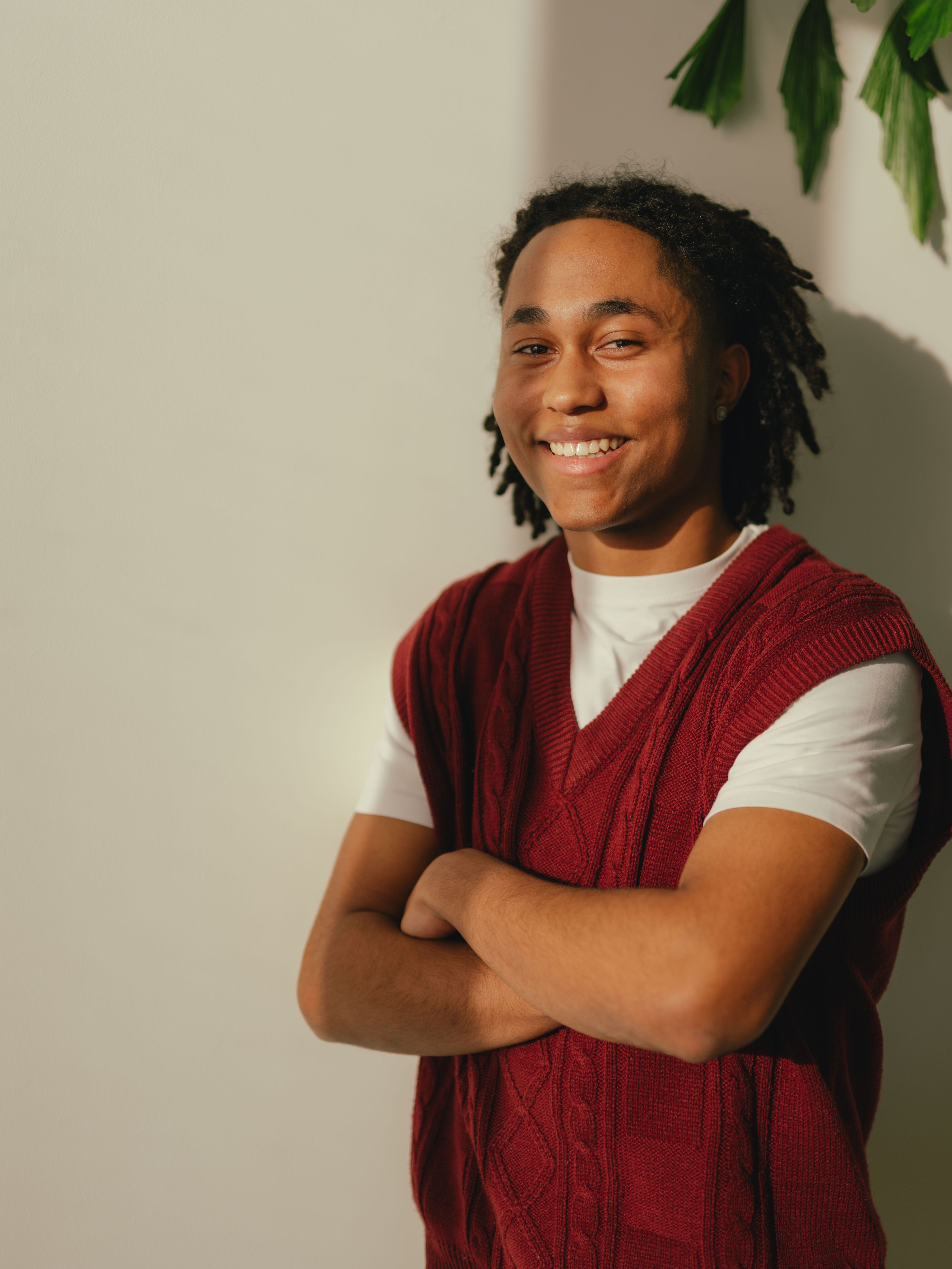 Una persona è in piedi davanti a una parete chiara con le braccia incrociate davanti al corpo. Indossa una t-shirt bianca sotto un capo lavorato a maglia e senza maniche, di colore rosso scuro. Sul lato destro dell’immagine spuntano diverse foglie verdi.