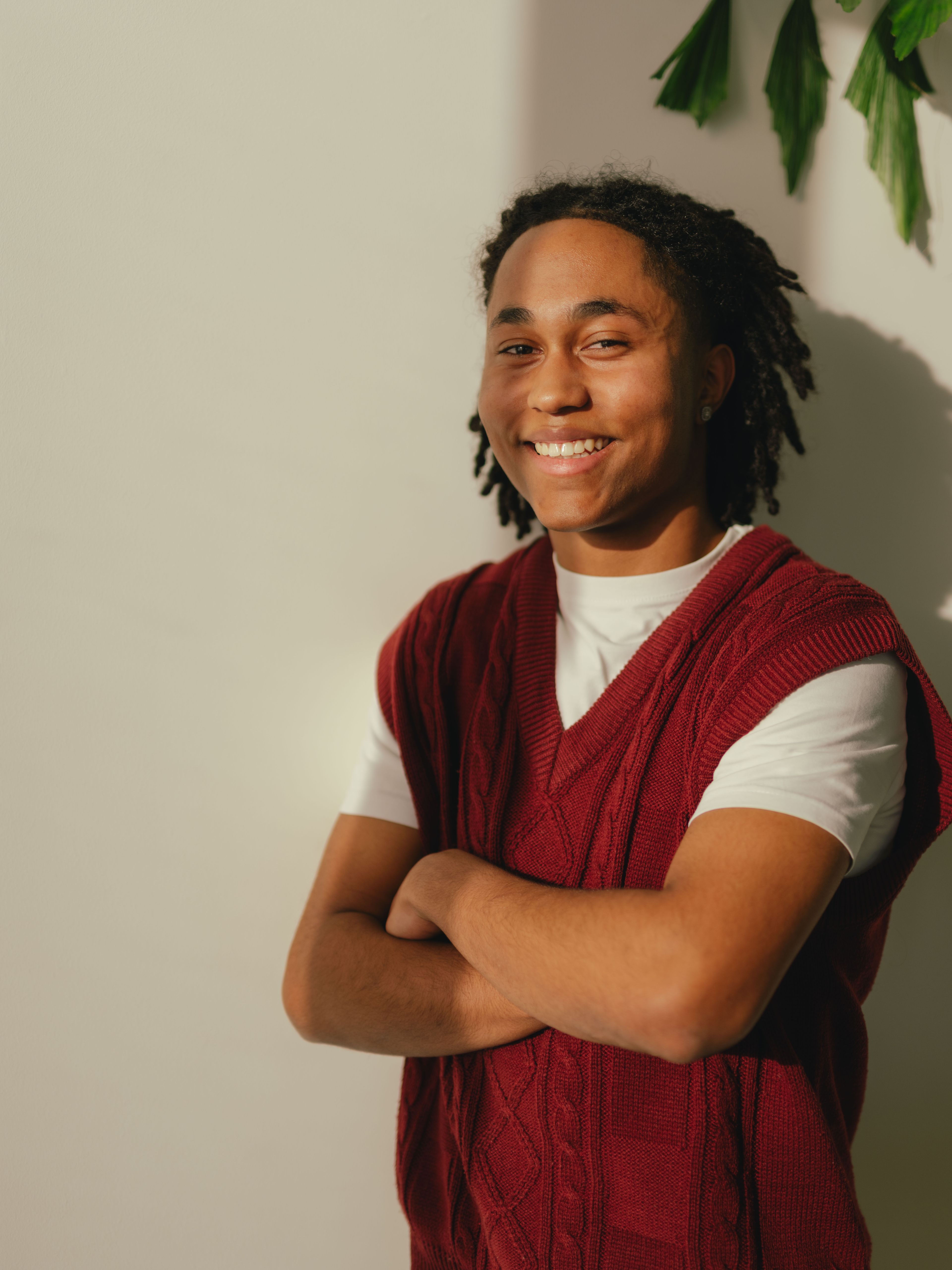 A person is standing in front of a light wall with their arms folded in front of their body. They are wearing a white T‑shirt under a dark red, knitted, sleeveless top. Several green plant leaves protrude into the picture on the right-hand side.
