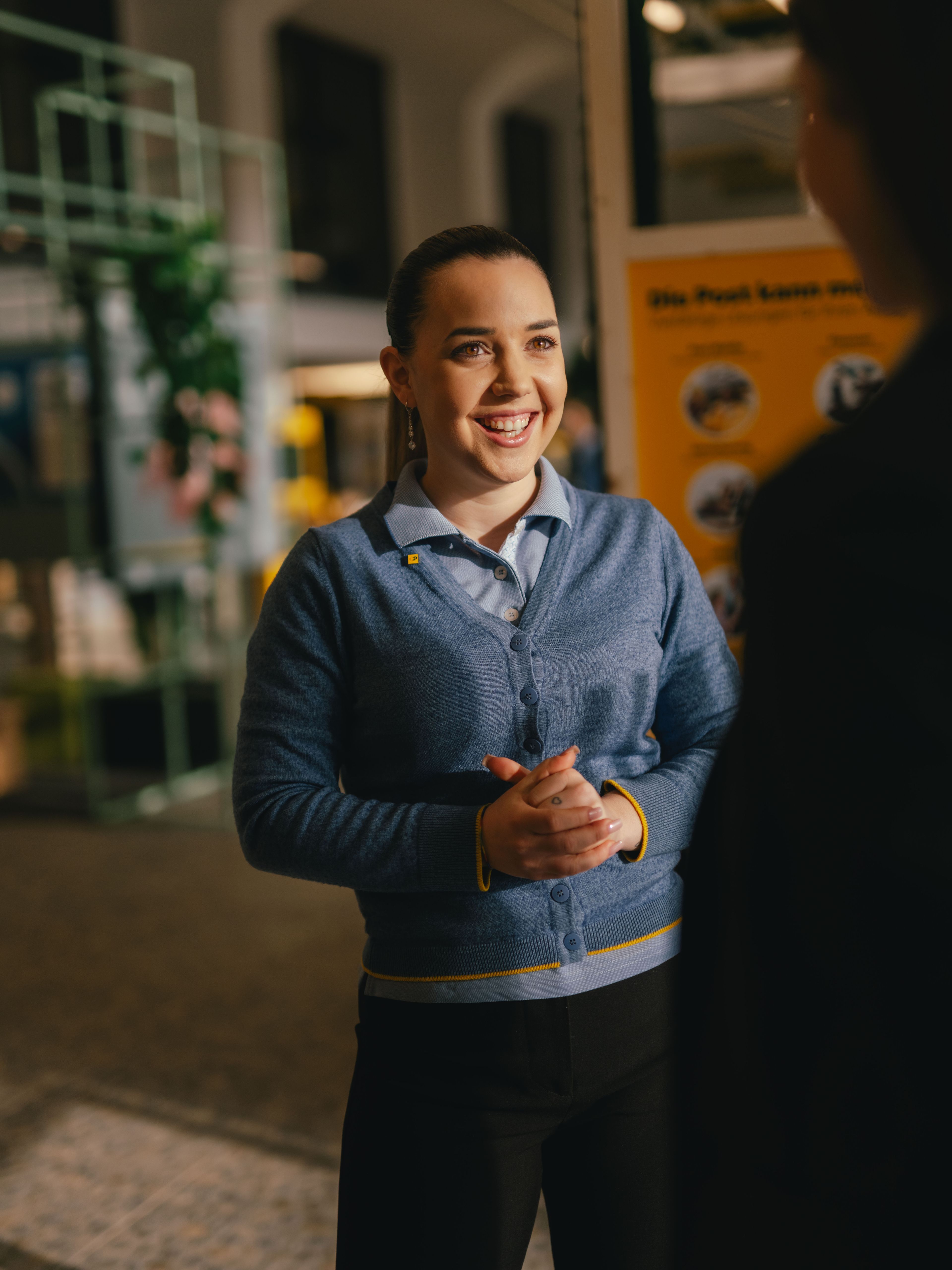 A person stands indoors and is engaged in conversation with another person in the foreground. The person is wearing a blue cardigan over a shirt and has hands clasped in front. In the background, informational displays and exhibition structures are visible.