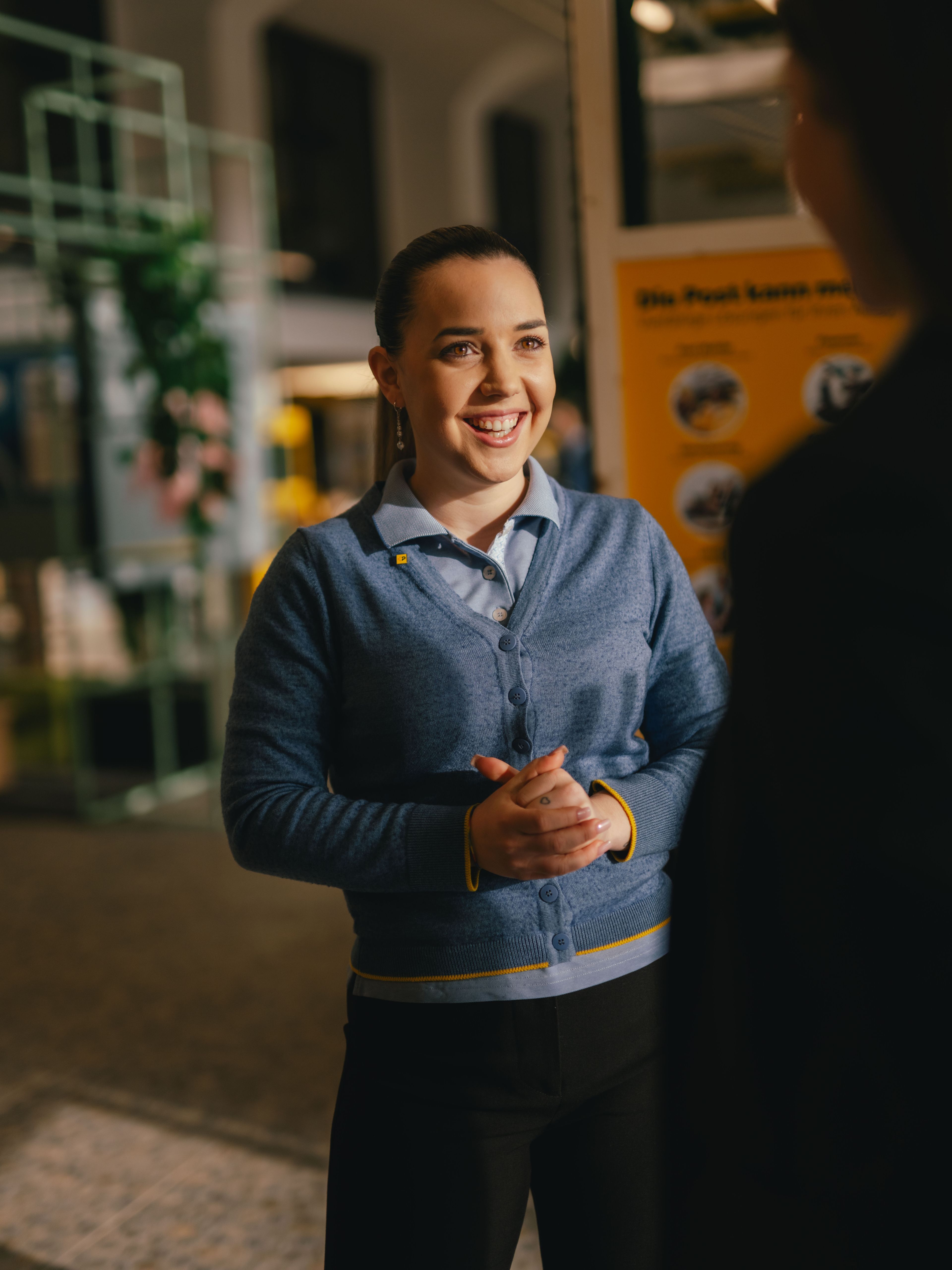 Une personne se tient à l’intérieur et s’adresse à une autre personne au premier plan. La personne porte un cardigan bleu par-dessus une chemise et garde les mains jointes devant elle. À l’arrière-plan se trouvent des panneaux d’information et des structures d’exposition.